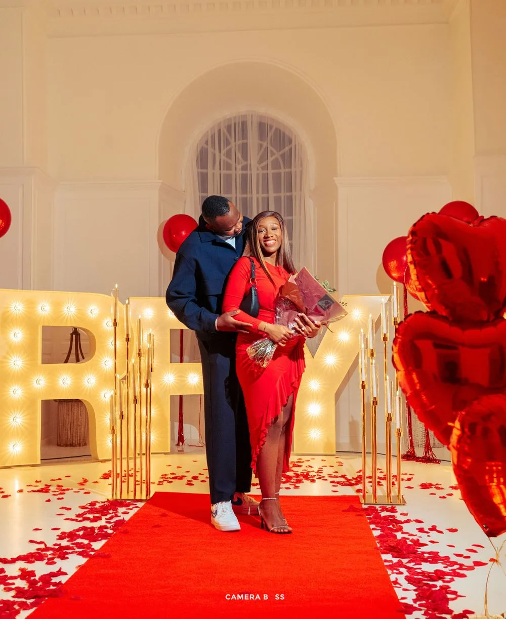 A couple shares a romantic moment on a red carpet surrounded by illuminated letters and rose petals in an elegant setting.