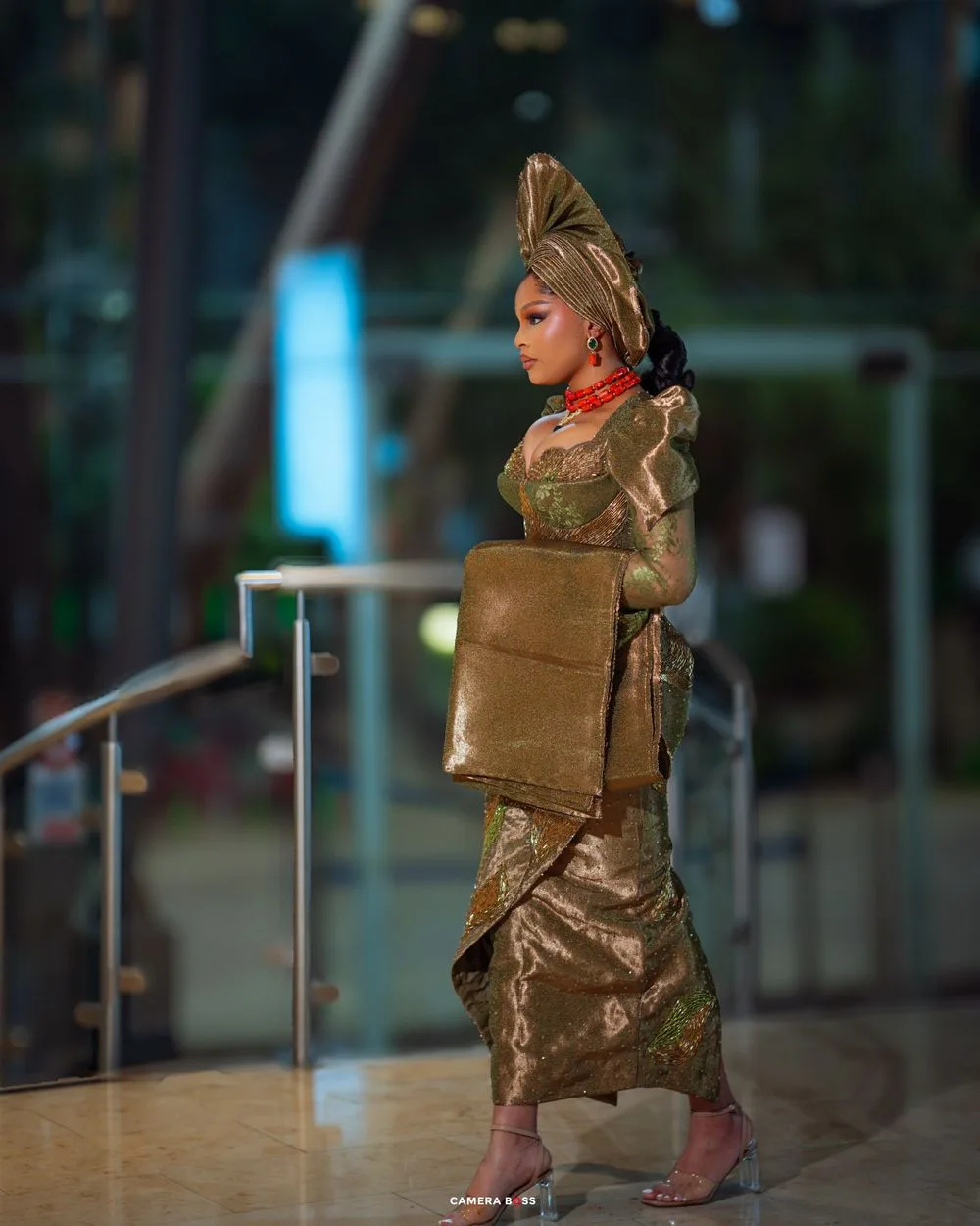 A side view of a model in an elegant gold dress and matching headpiece posing near a glass railing.