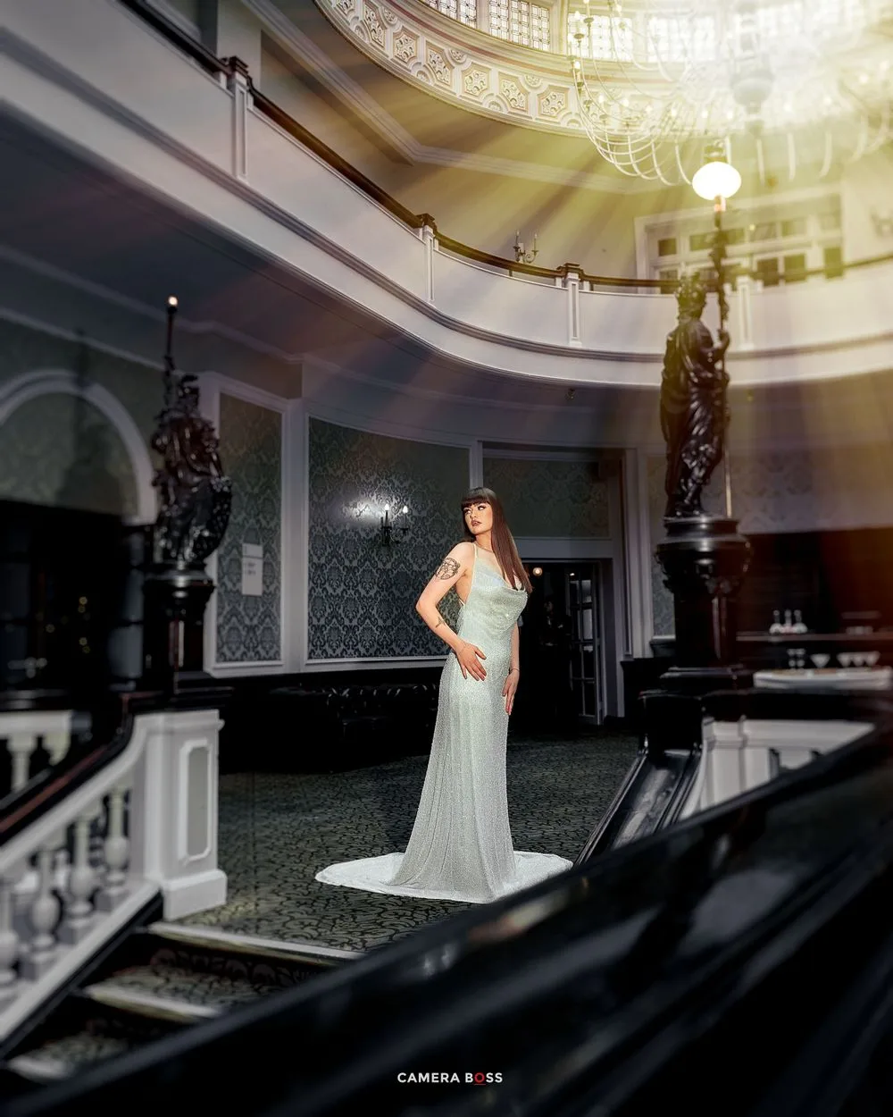 A woman in a silver gown stands on elegant stairs beneath a domed ceiling with dramatic lighting.