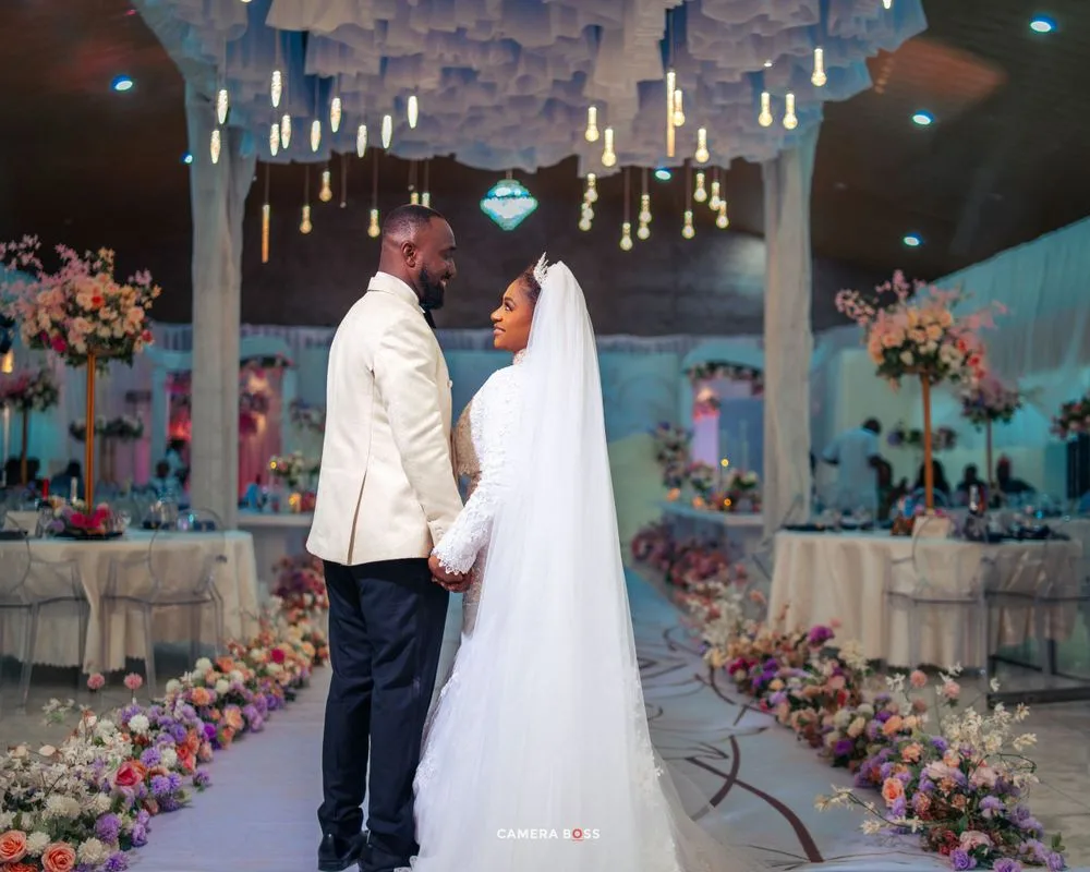 A couple at their wedding ceremony under a canopy decorated with hanging lights and colorful floral arrangements.