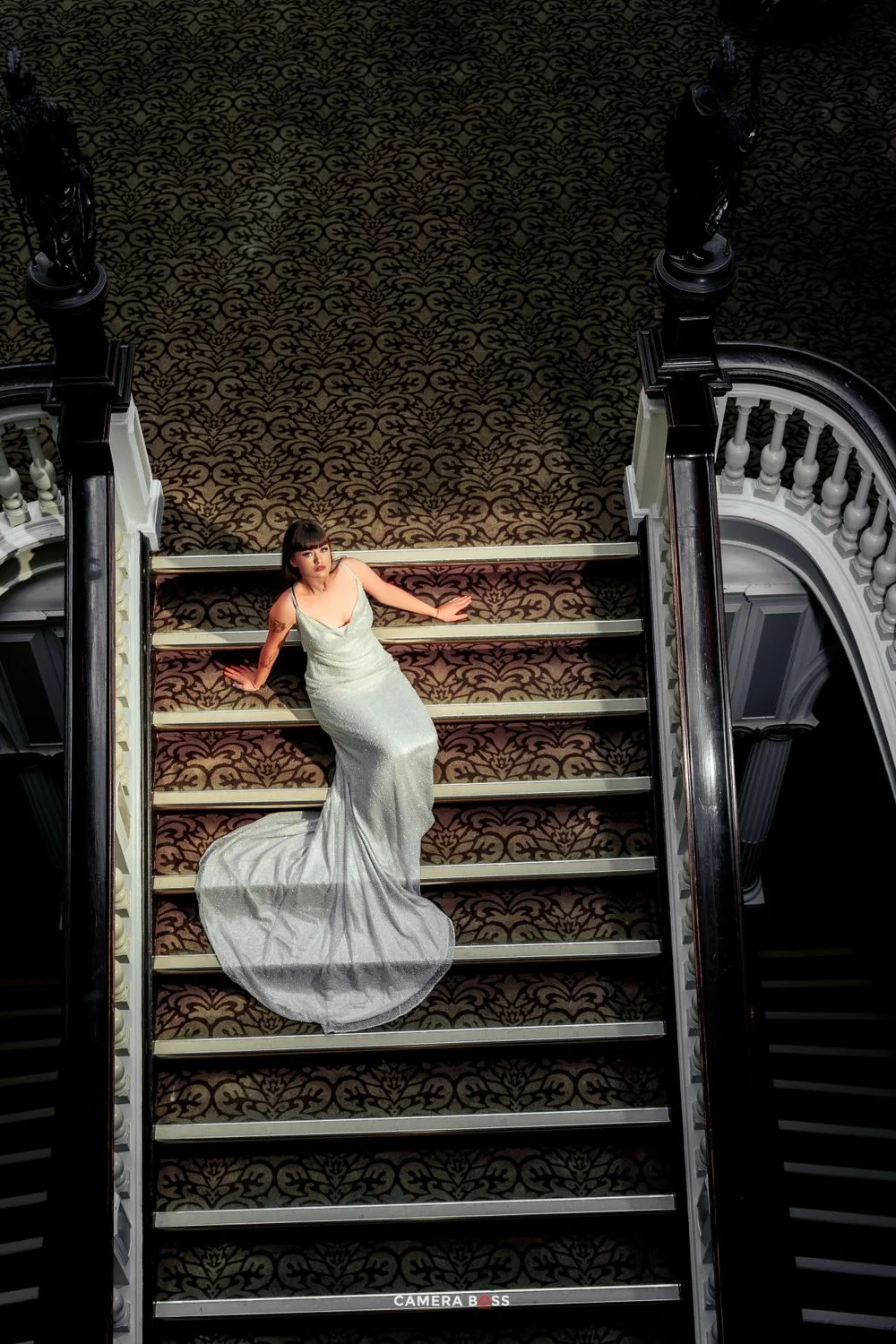Elegant figure in flowing silver gown descends ornate carpeted staircase with decorative balustrade in dramatic lighting.