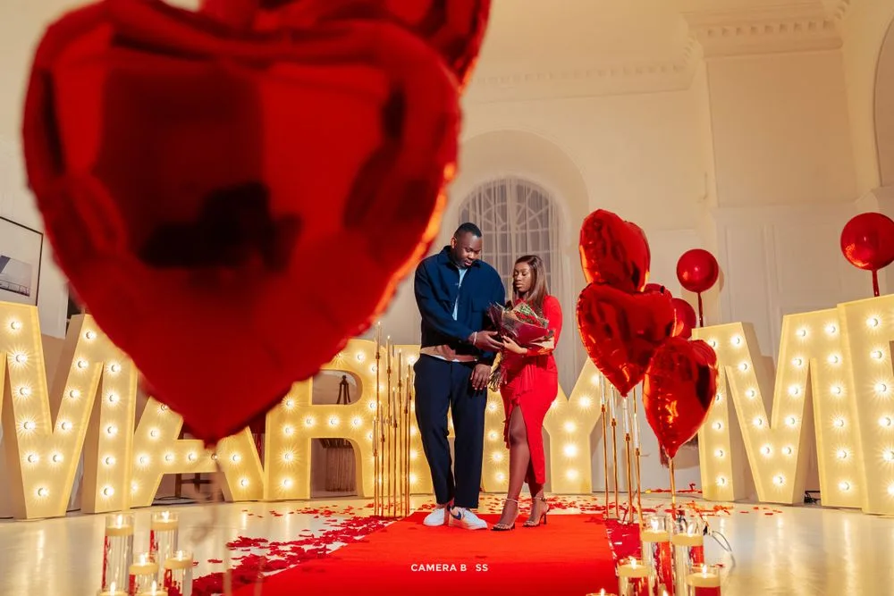 A romantic proposal setup with red heart balloons and illuminated 'MARRY ME' letters along a red carpet walkway.