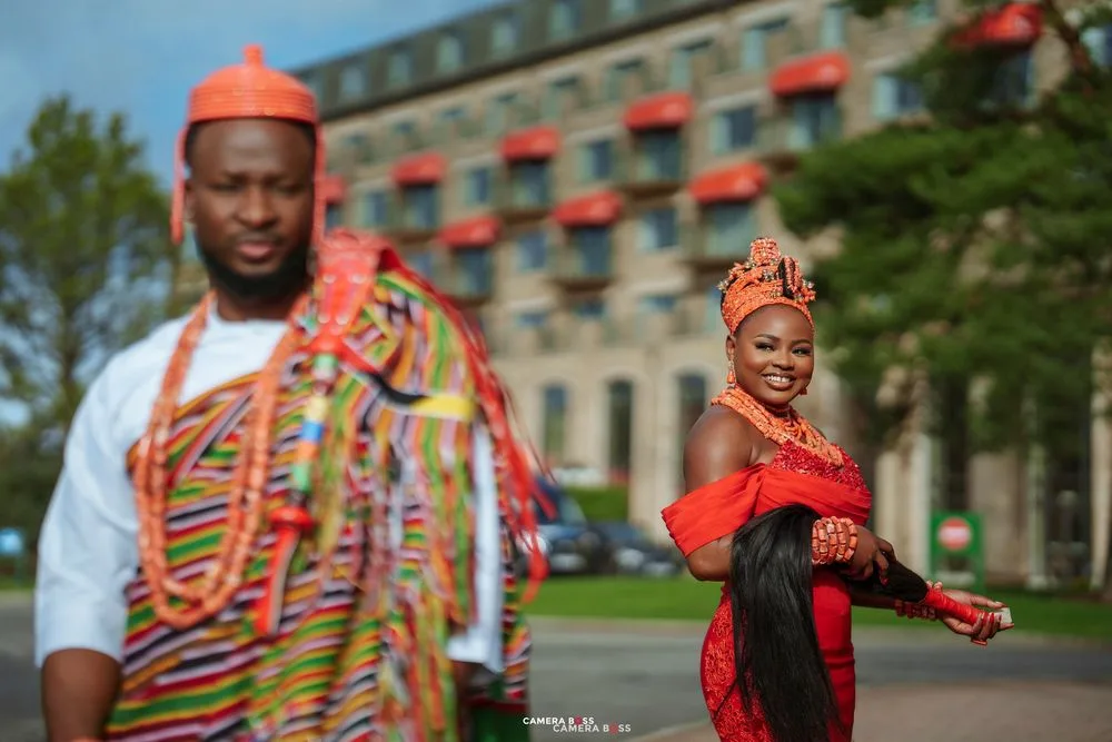 Couple in vibrant African traditional attire posing outdoors, woman in red and man in colorful kente fabric.