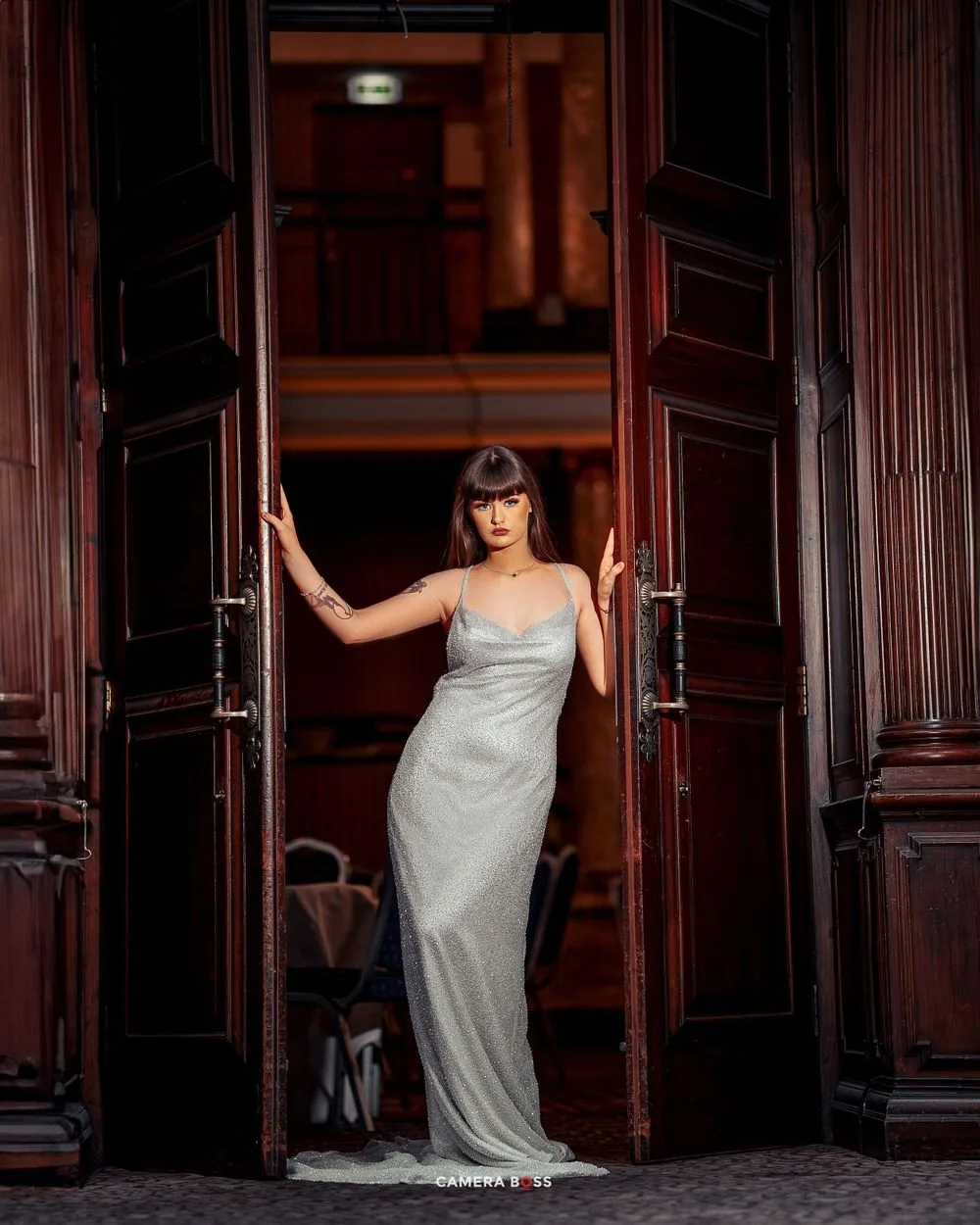 A woman in a silver evening gown poses between ornate wooden doors in a dramatic entrance.