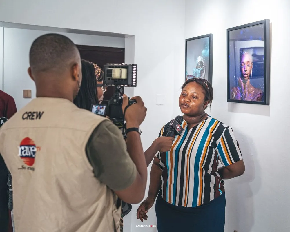 A TV crew member in a beige vest films someone speaking in front of framed portrait photographs on a wall.