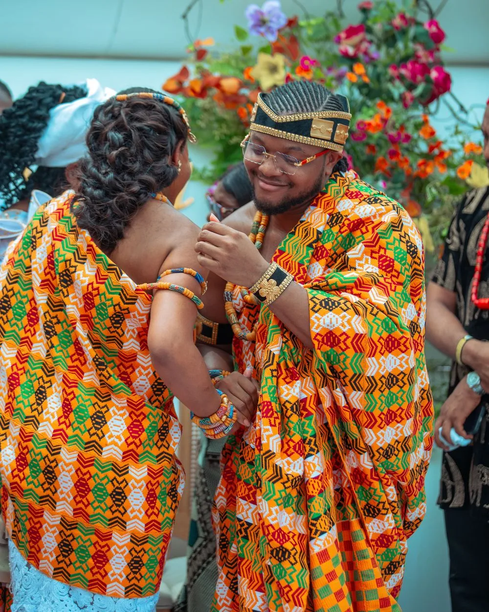 Two people wearing matching vibrant orange and green patterned traditional African kente cloth outfits dancing together at an event.