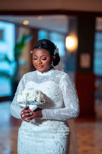 A bride in an elegant long-sleeved white lace wedding gown holds a white rose bouquet in an indoor venue.