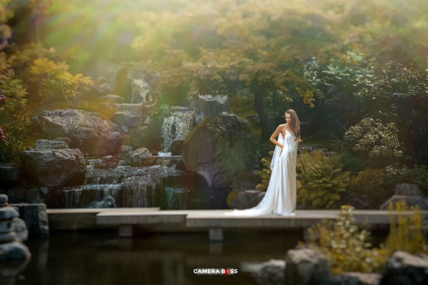 White-gowned figure stands on wooden dock by tranquil waterfall in ethereal garden setting at golden hour.