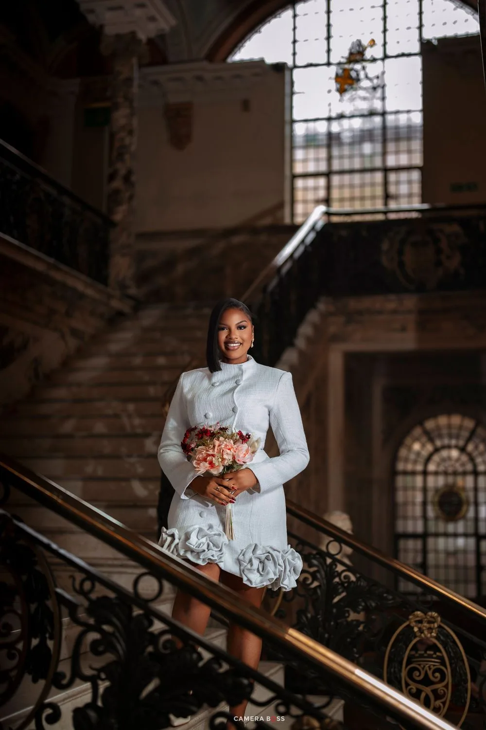 Bride in white short wedding dress holding floral bouquet on ornate staircase in elegant historic venue.