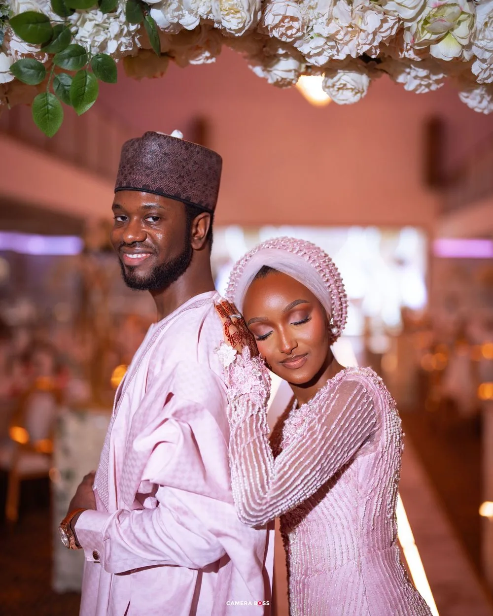 Romantic couple poses in matching white traditional attire during indoor wedding photoshoot with warm lighting.
