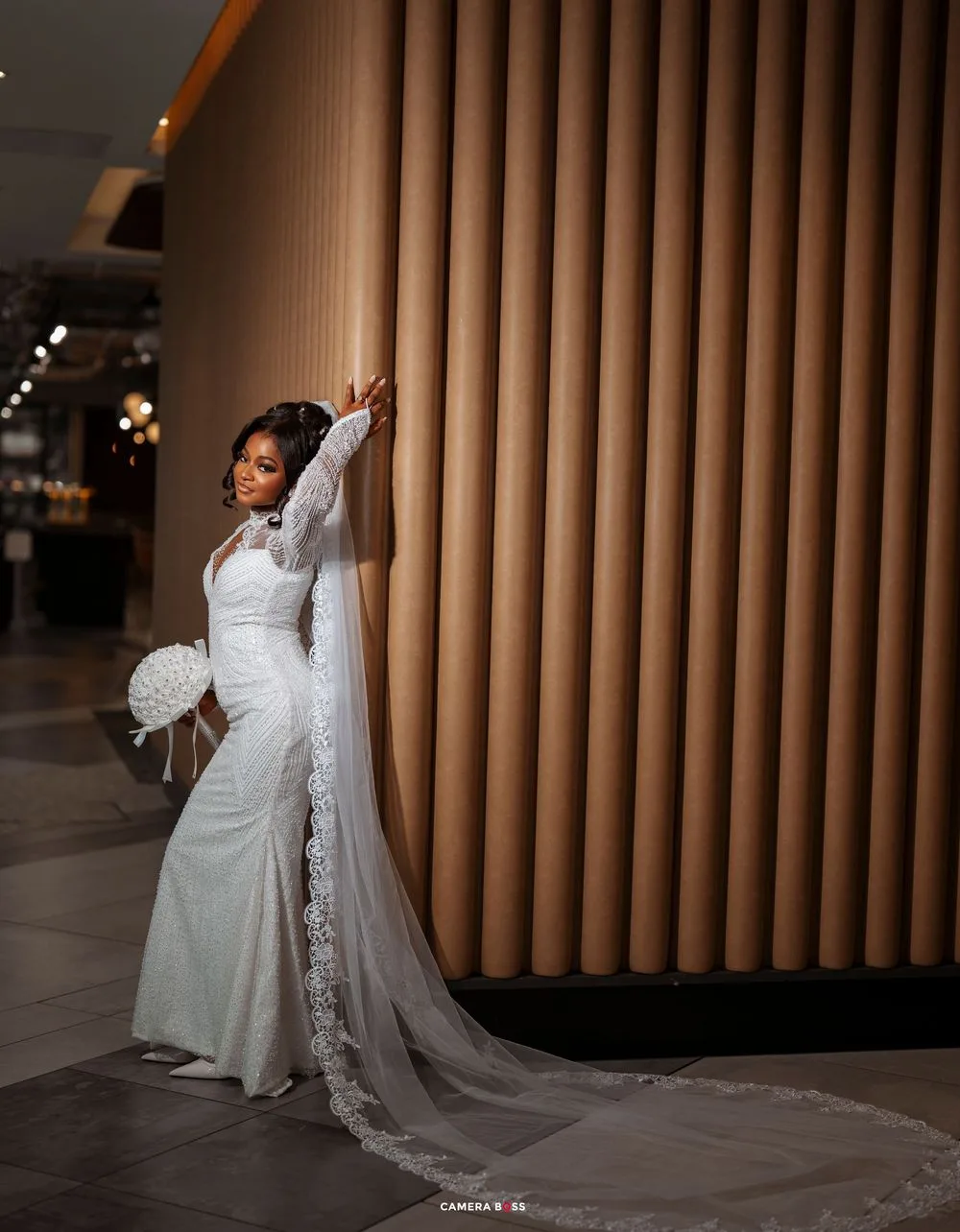 A bride in an elegant white lace wedding gown with a long veil poses dramatically against brown architectural columns.