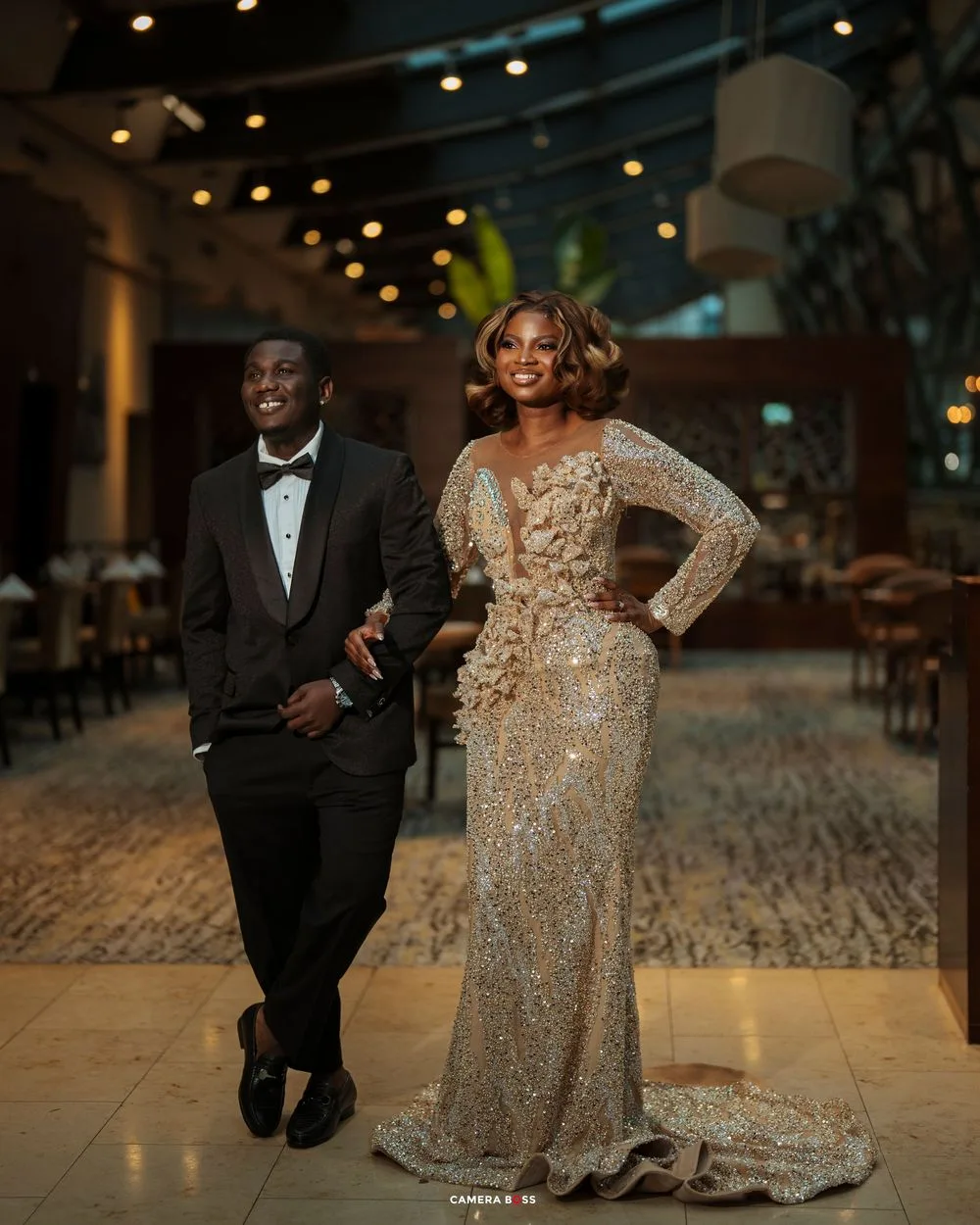 Elegant couple posing in formal attire, man in black tuxedo and woman in gold sequined gown, at upscale venue.