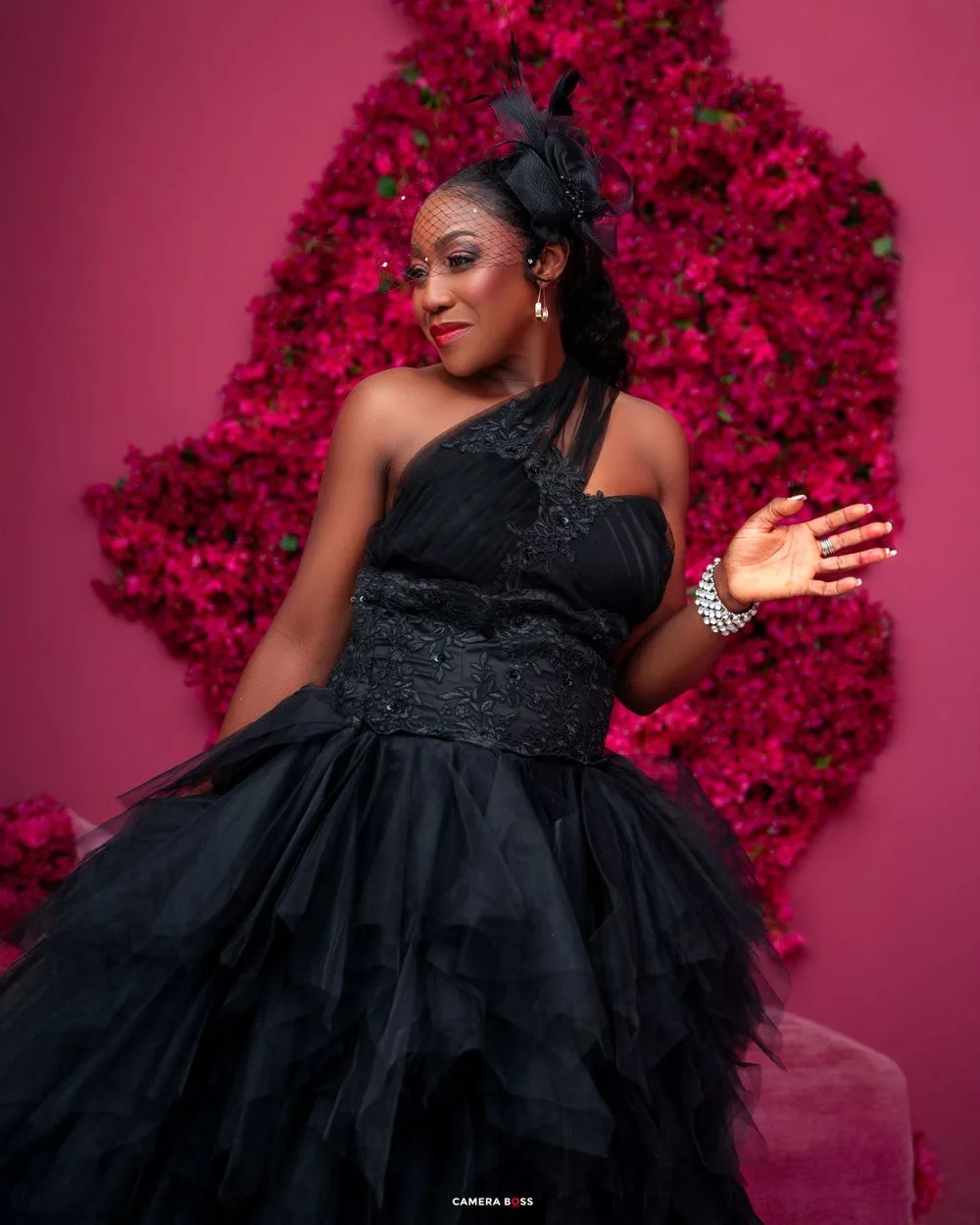 A figure in an elegant black one-shoulder tulle gown poses against a vibrant pink floral backdrop.
