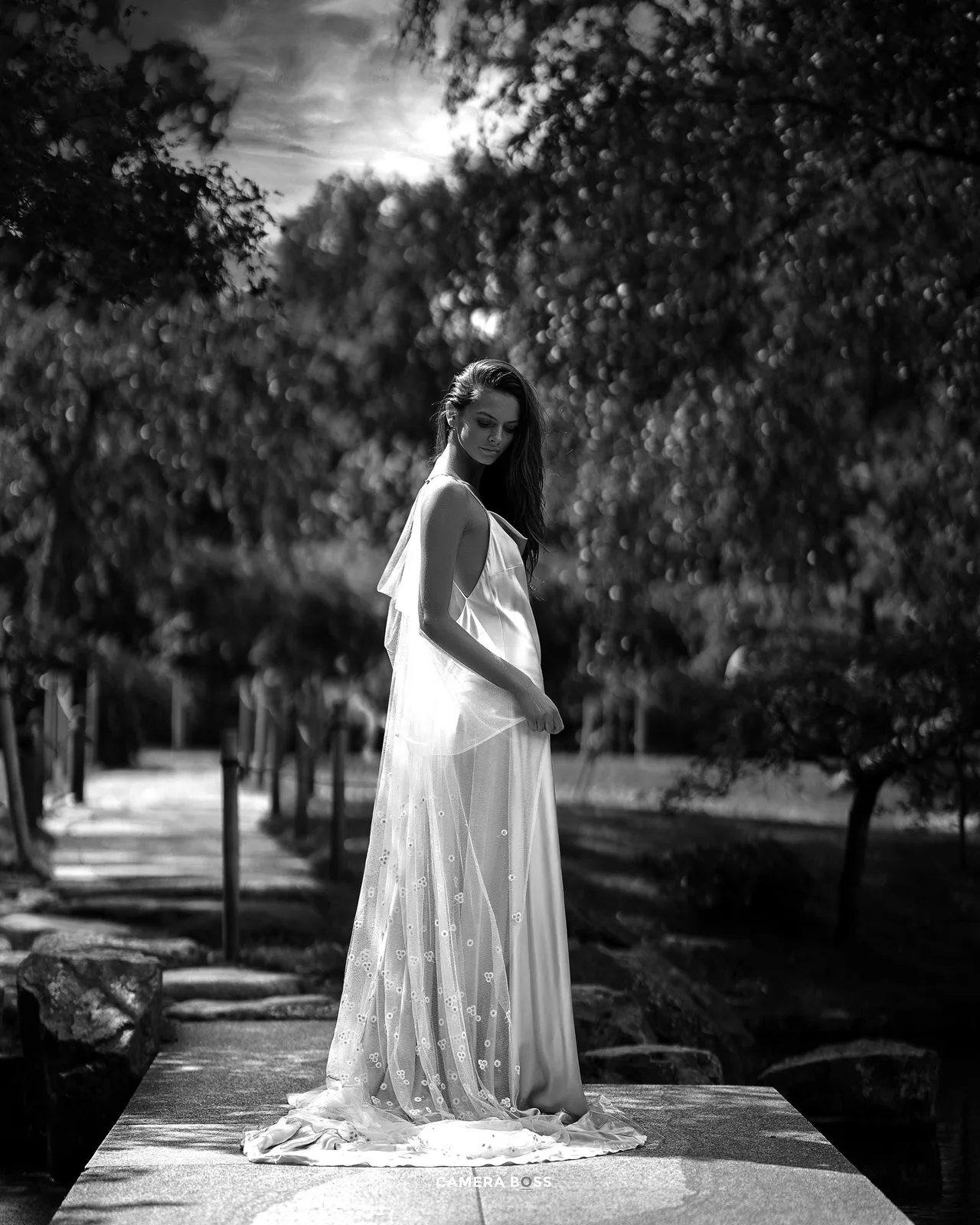 A black and white portrait of someone in an elegant white dress standing on a stone surface in a park setting.