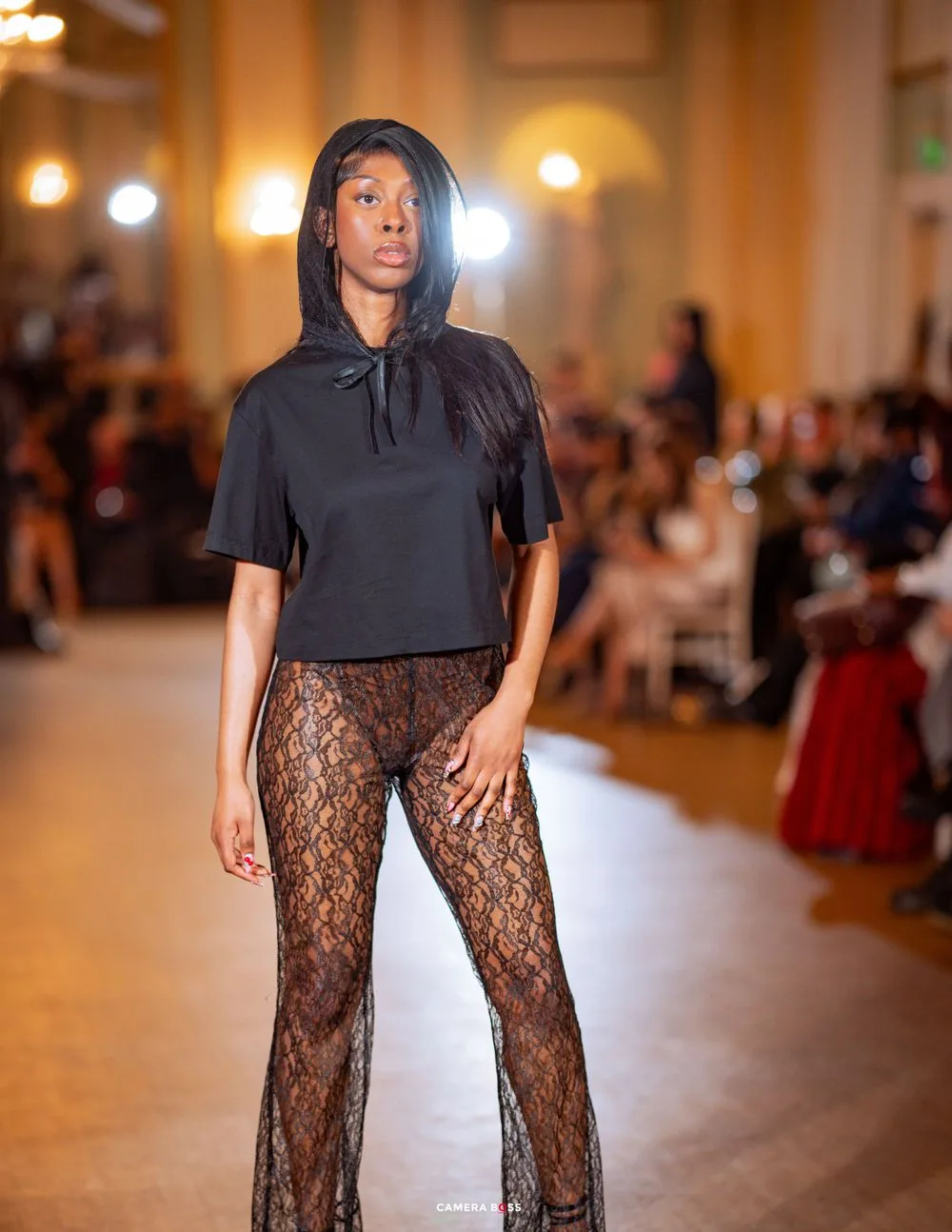 Model presents black hooded top with sheer lace pants on illuminated runway in grand ballroom.