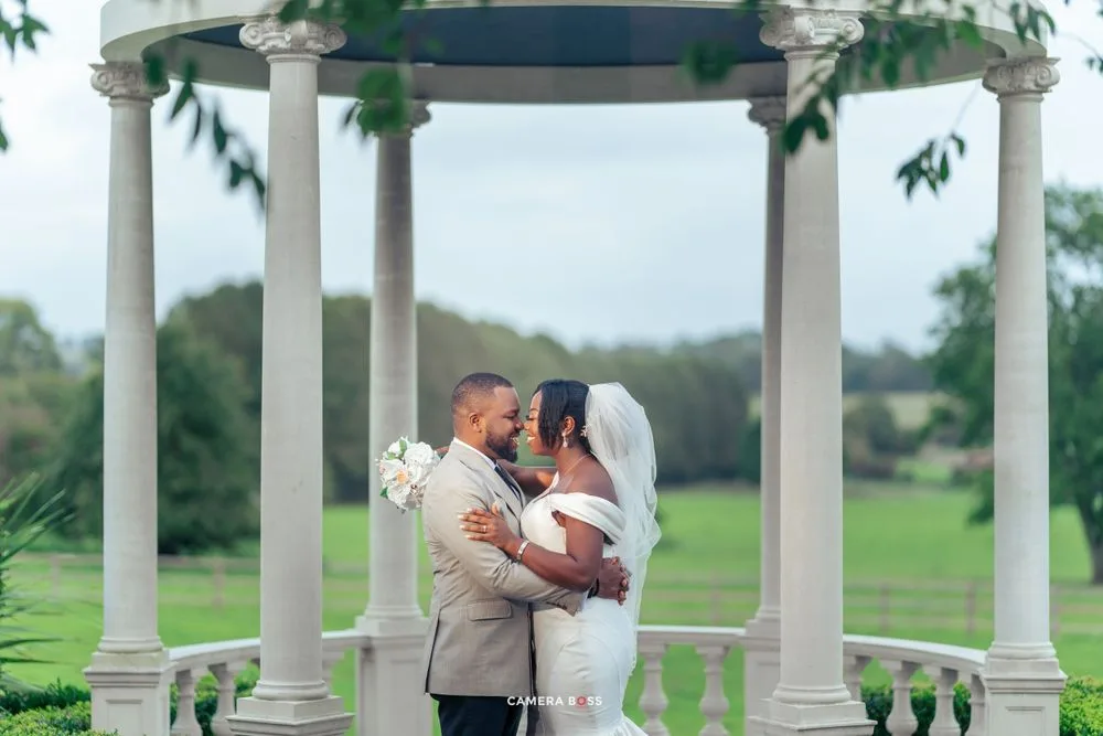 A couple shares a romantic moment under an elegant white gazebo surrounded by lush green landscaping.