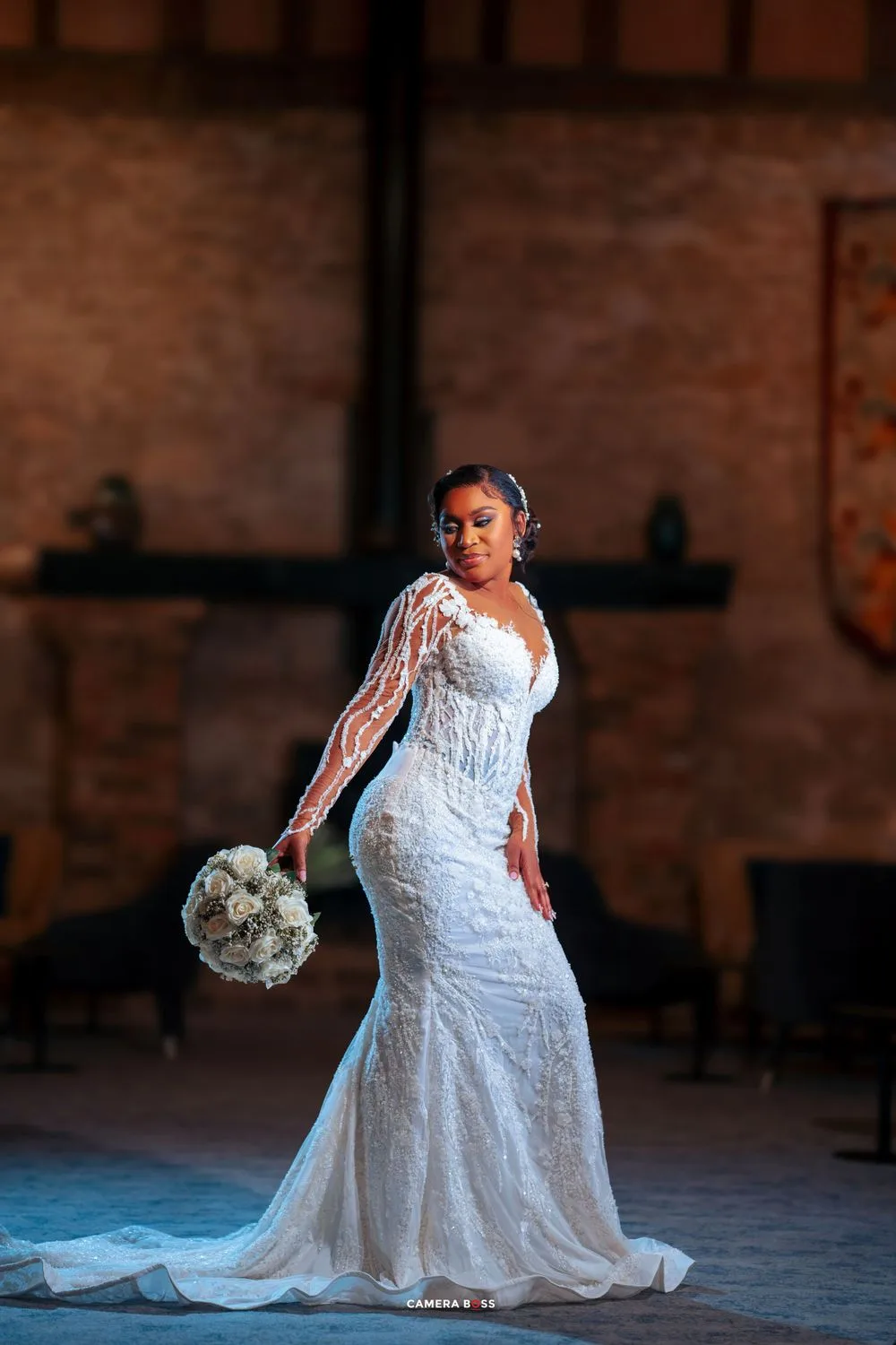 An elegant bride wearing a fitted lace wedding dress stands gracefully with white flowers against a rustic brick wall.