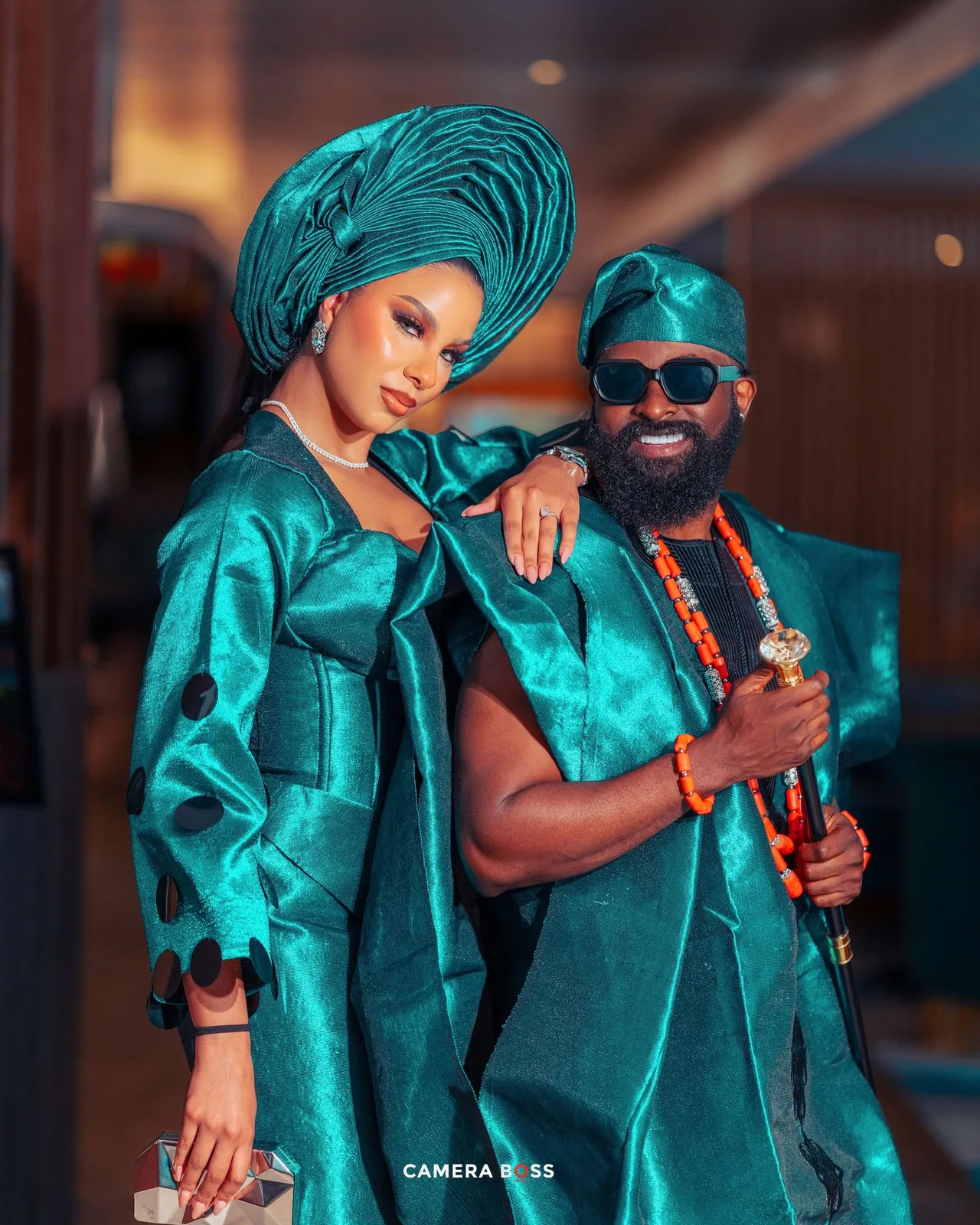 A couple in matching turquoise traditional African attire poses together against a blurred background.