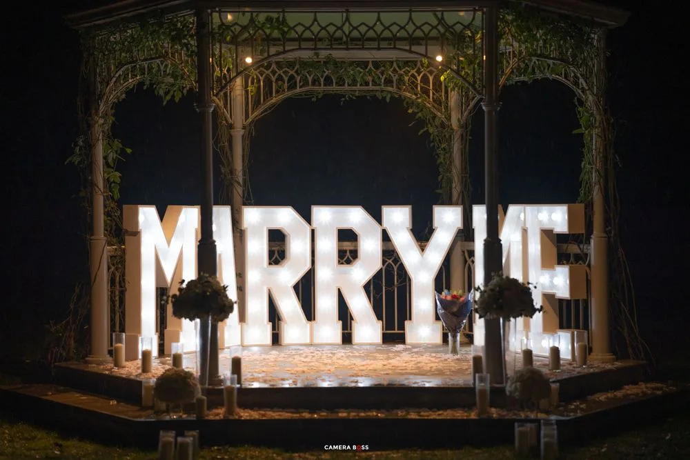 Illuminated 'Marry Me' proposal sign glows on a vintage ornate stage with decorative iron arches and candles.