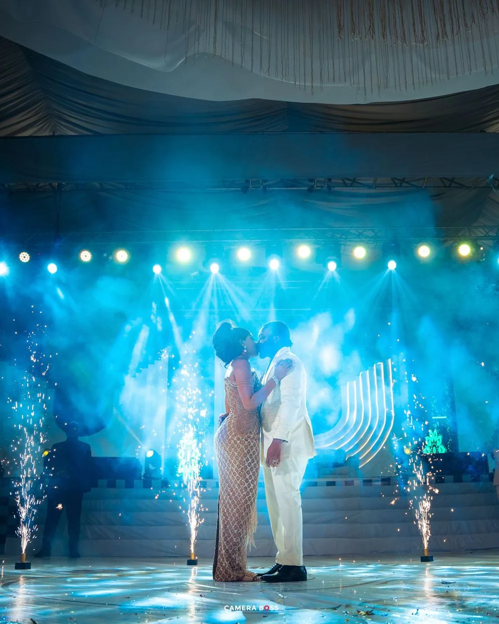 Couple dancing on an illuminated stage with blue lighting, pyrotechnics and draped fabric decor at an elegant event.