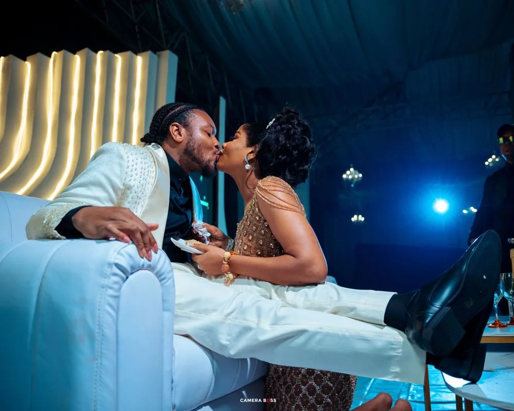 A couple shares an intimate moment while sitting on white furniture at an evening celebration with neon lighting.