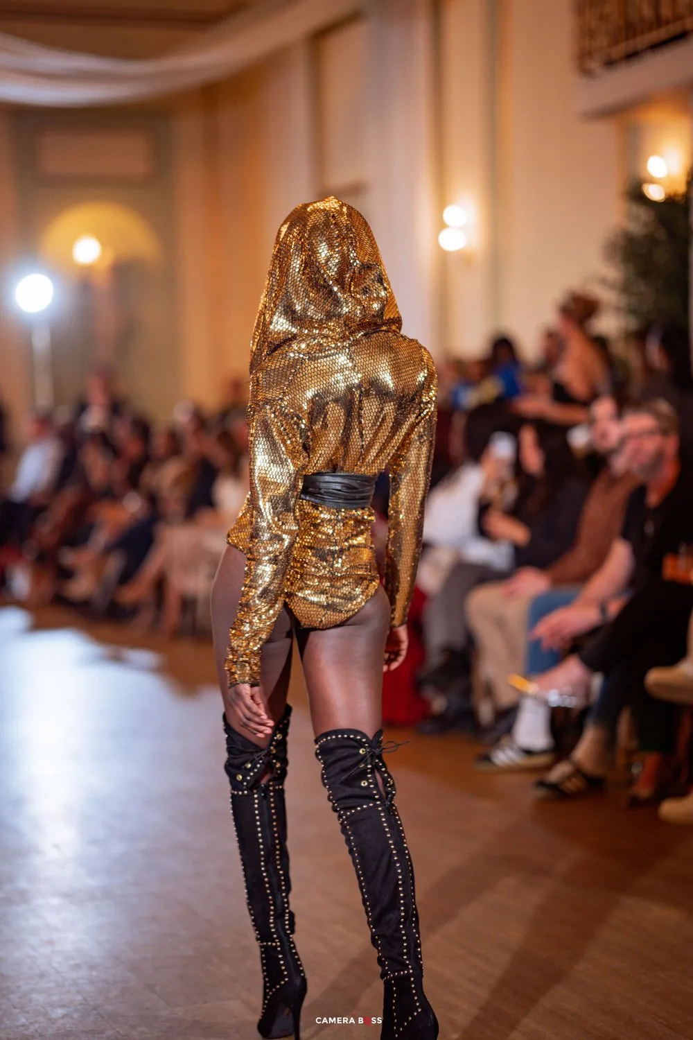 Back view of model wearing shimmering gold hooded ensemble with black boots on fashion runway.