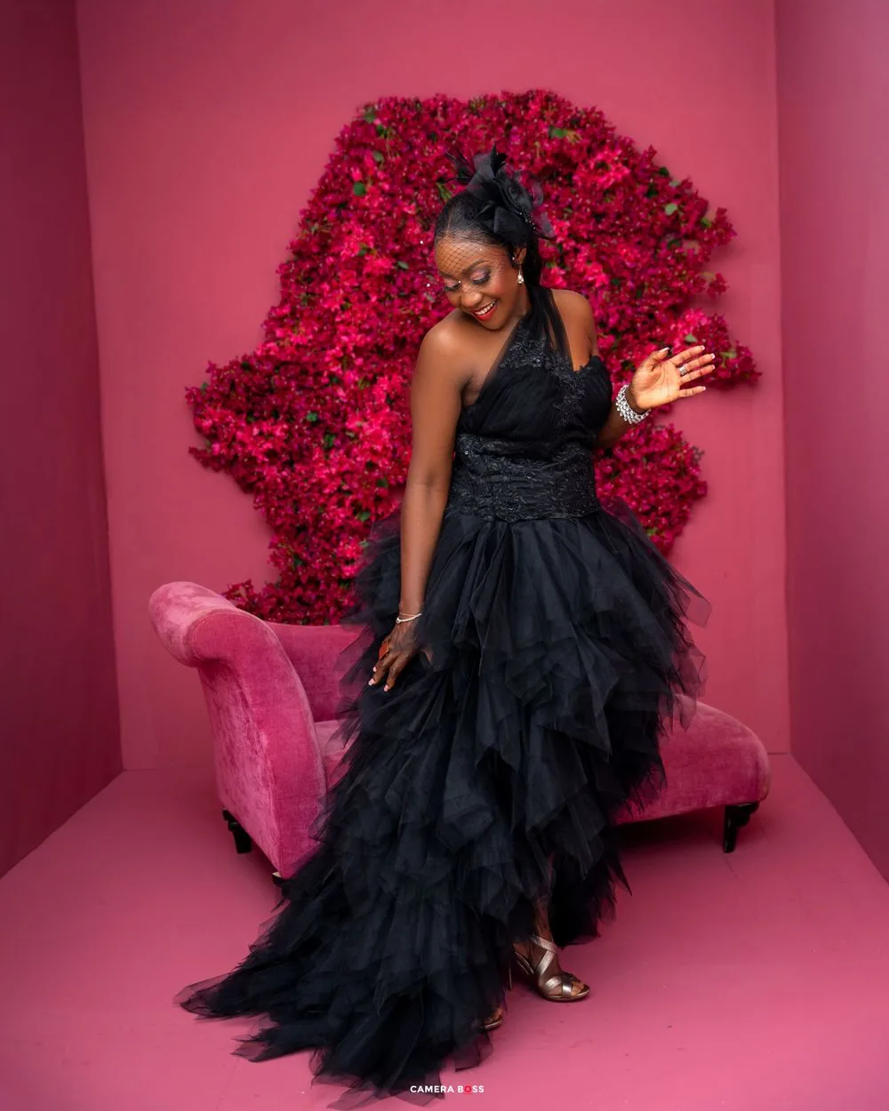 Elegant black tulle gown stands out against vibrant red floral backdrop and pink chaise lounge in studio setting.