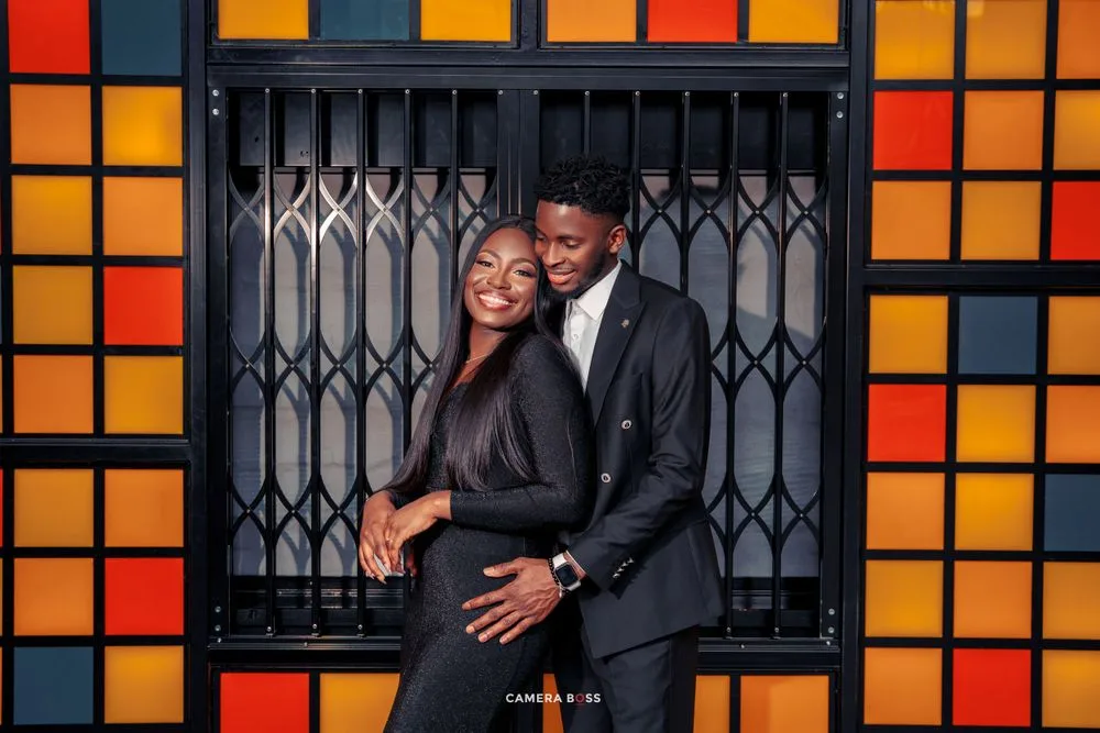 A stylish couple embraces in front of a colorful geometric wall with orange and red squares and metal grating.