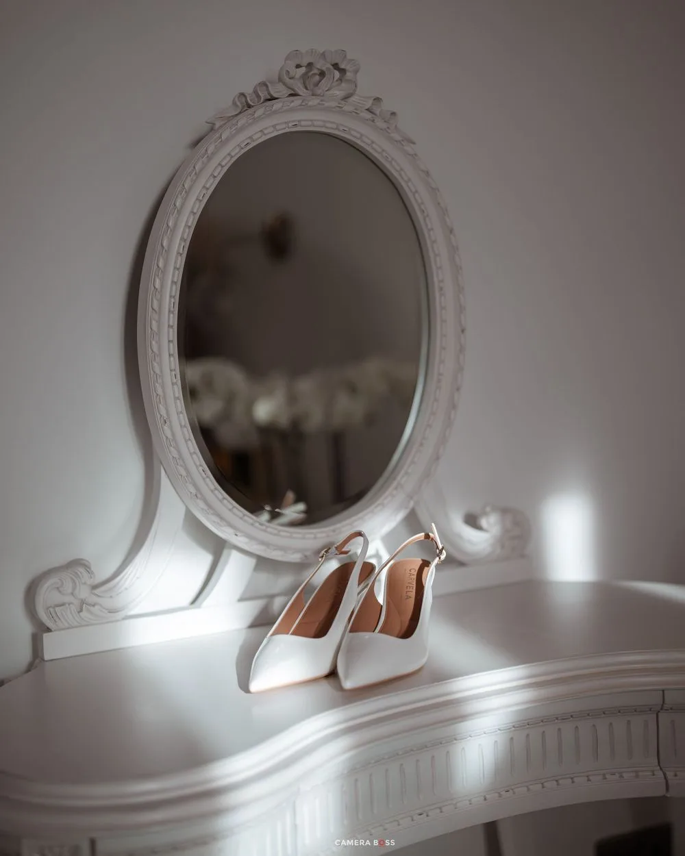 Elegant white bridal heels displayed on vintage white vanity table with ornate oval mirror.
