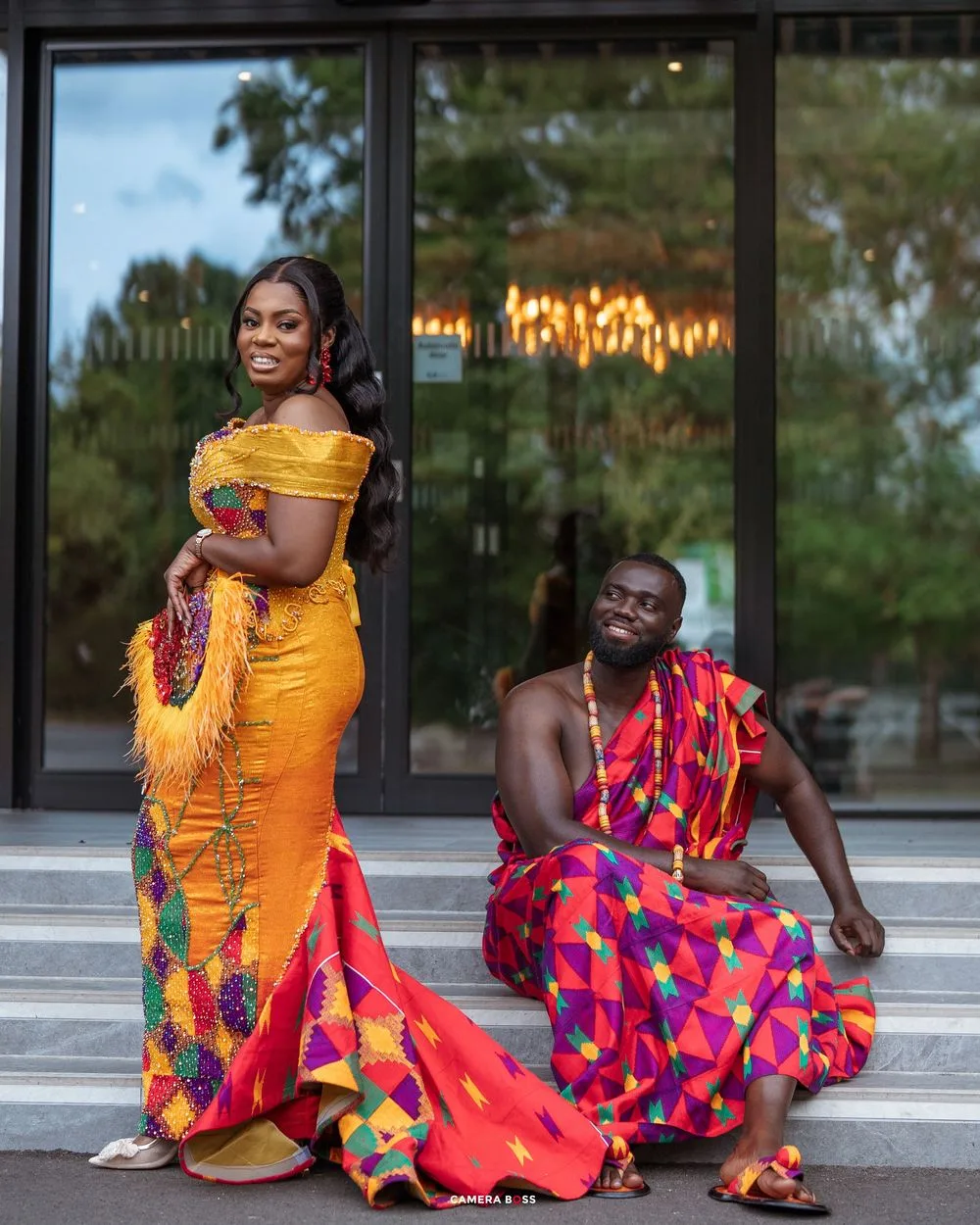 Couple in vibrant Kente cloth traditional Ghanaian attire, woman in yellow mermaid gown, man seated in colorful fabric.