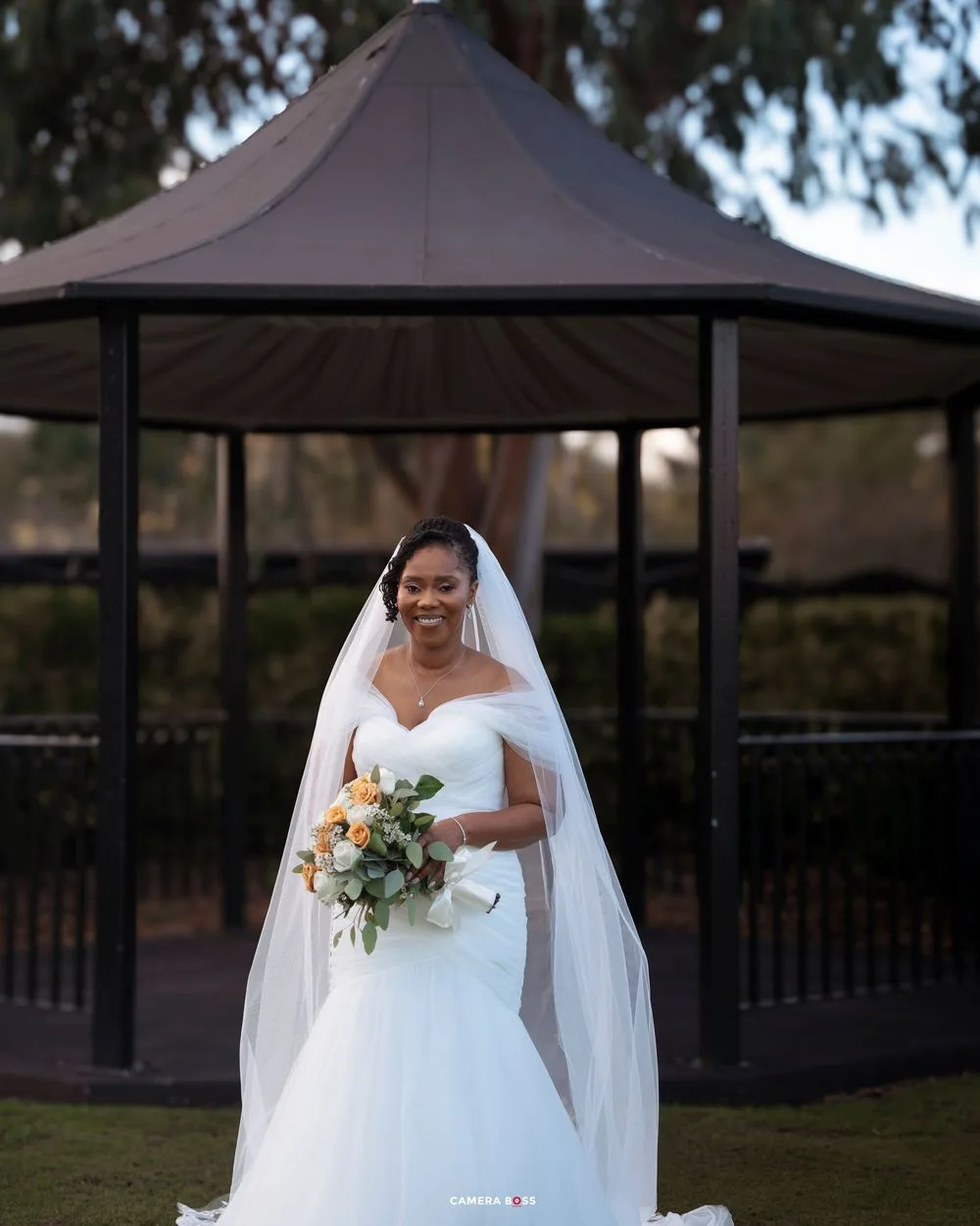 Smiling bride in white mermaid gown and veil holds bouquet before elegant dark gazebo outdoors.