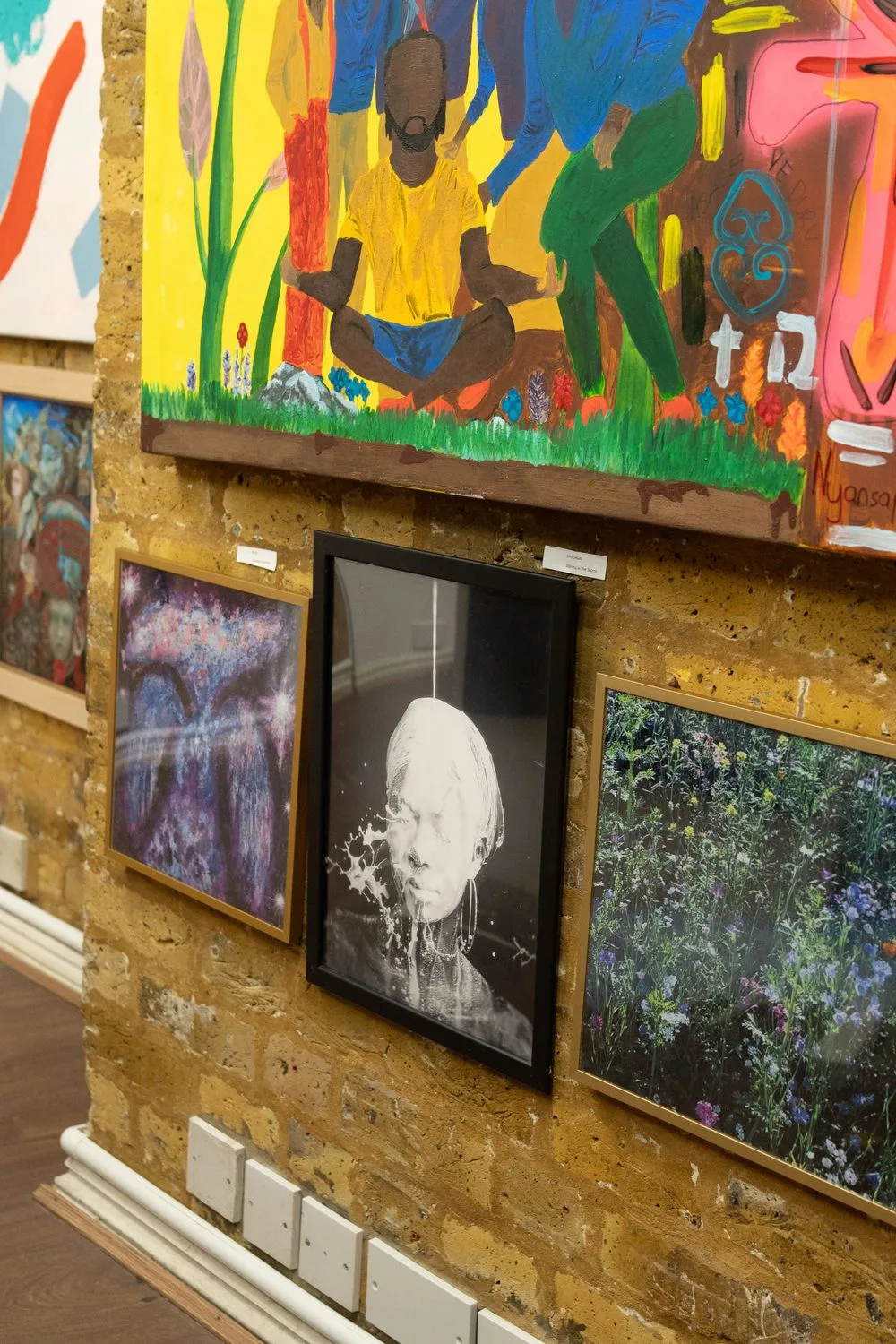 Colorful artwork display on a rustic yellow wall showing paintings with vibrant hues and contrasting images.