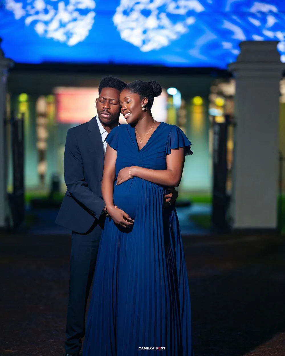 A couple in elegant evening attire share an intimate moment under blue lighting in an outdoor setting.