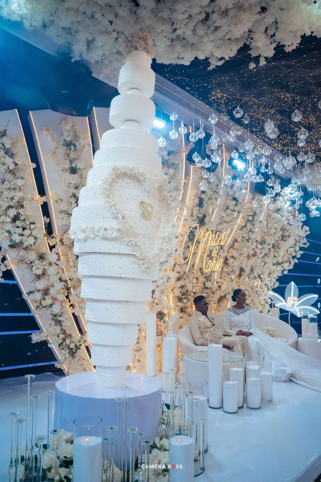 An elegant all-white wedding cake display with floral decorations and candles at a luxurious reception venue.
