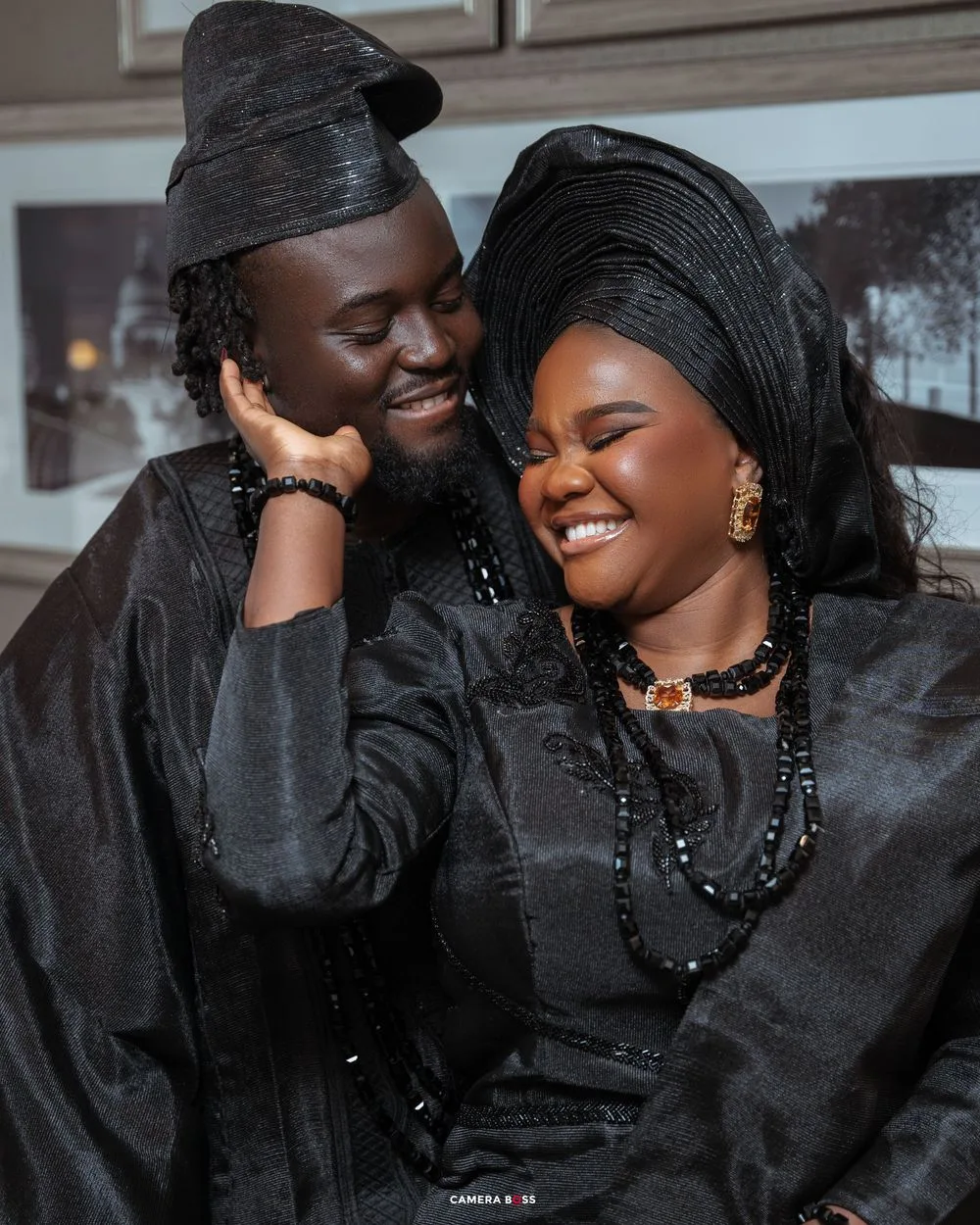 A couple in matching black traditional outfits and head wraps share a joyful moment while embracing and laughing together.