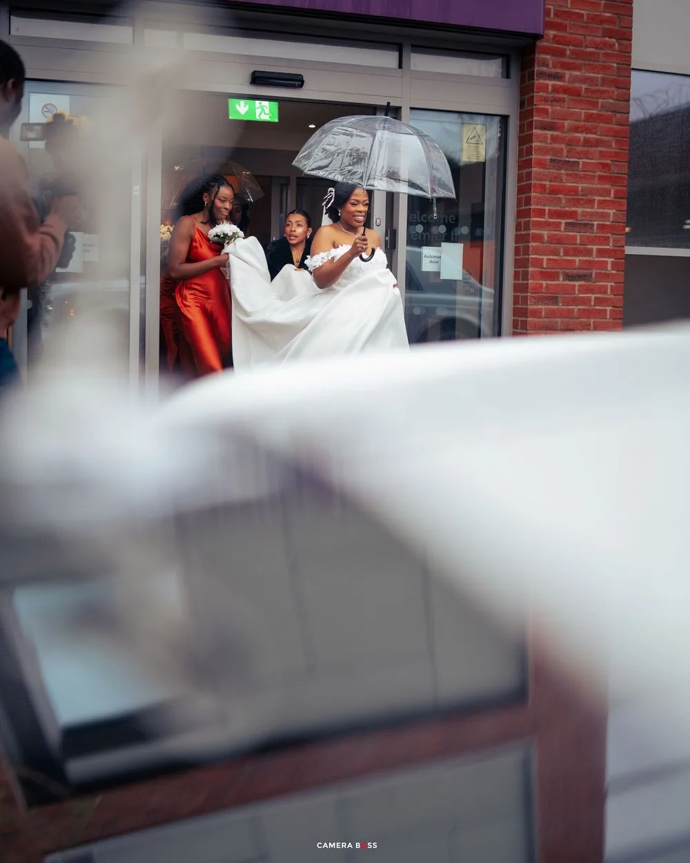 A bride in a white wedding dress exits a building while holding a clear umbrella during a rainy day.