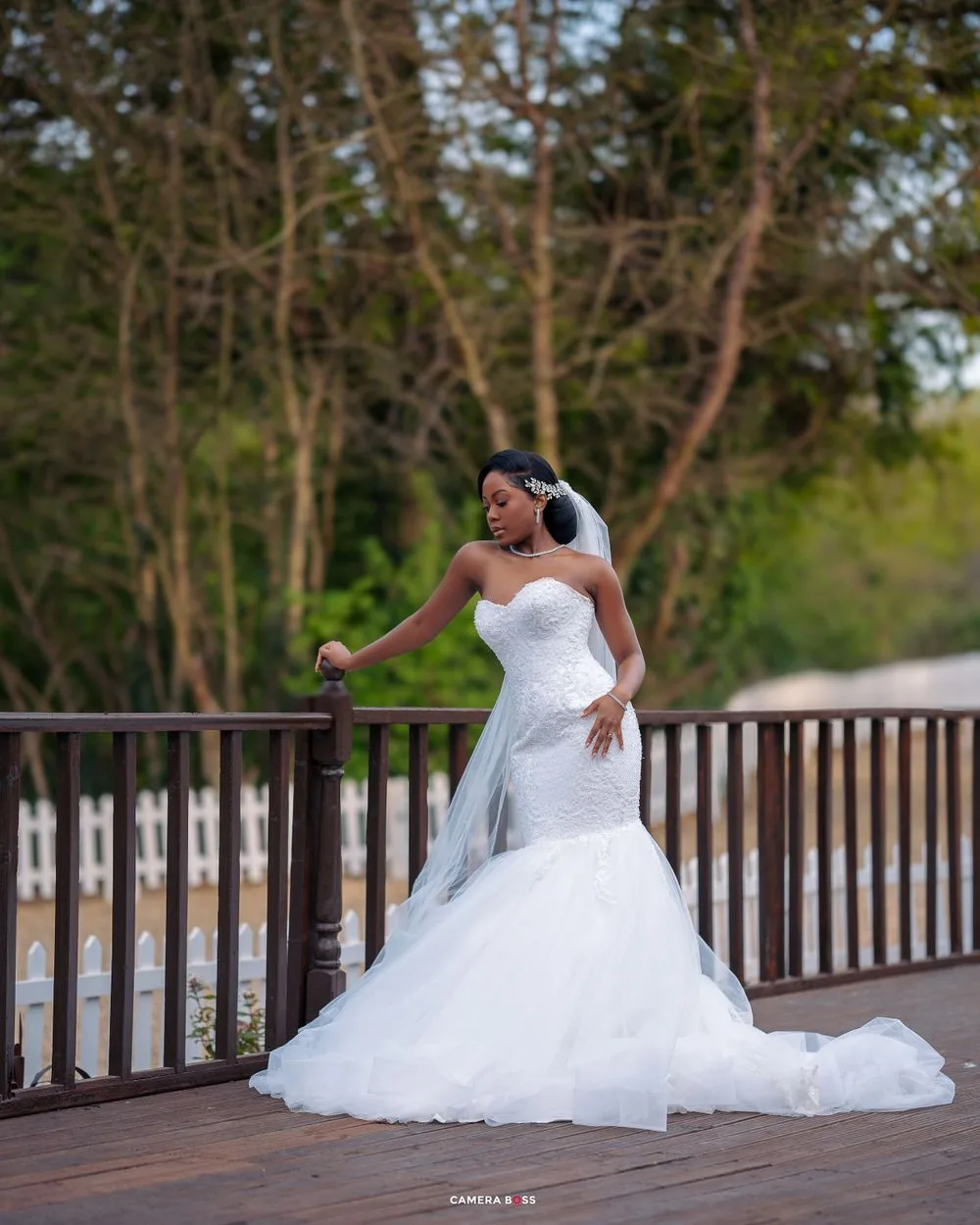 A bride in a stunning white mermaid wedding gown poses elegantly on a wooden deck surrounded by trees.
