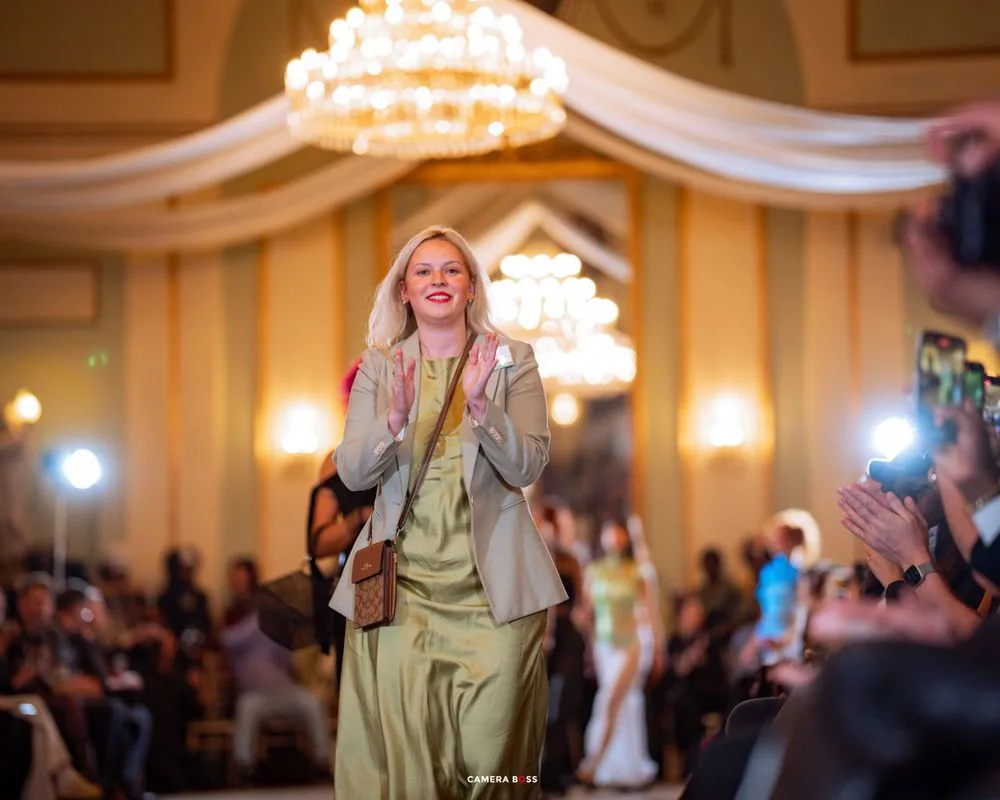 A smiling person walks down an elegant runway in a light green outfit as chandeliers glow overhead in a grand ballroom.