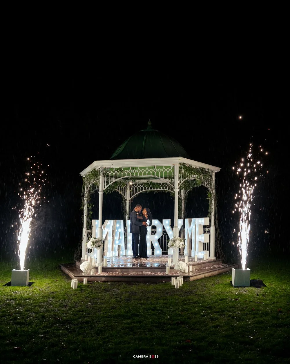 A white gazebo illuminated at night with sparklers and a lit-up MARRY ME sign for a romantic proposal.