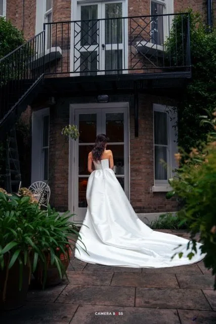 Elegant white wedding gown with dramatic cathedral train photographed from behind against a historic brick building entrance.