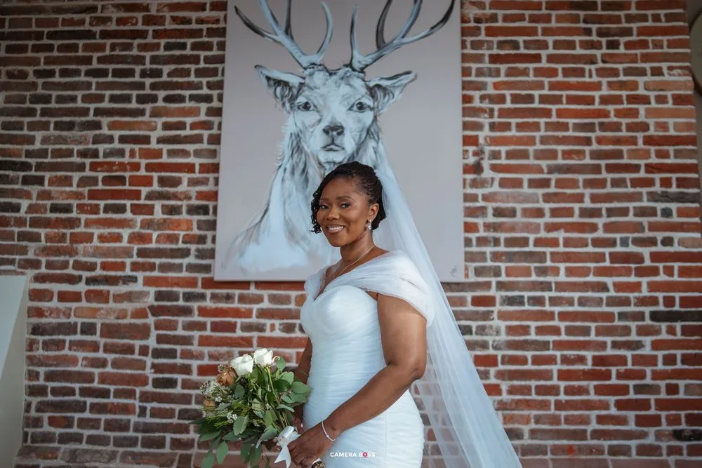 A bride in a white dress poses in front of a brick wall with a large deer artwork behind her.