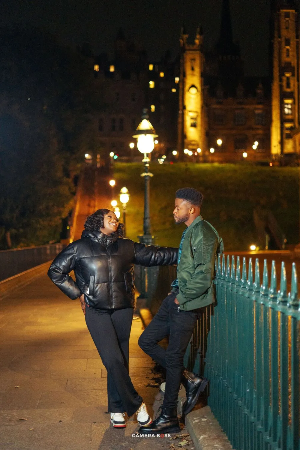 Two people share a conversation by railings at night with illuminated historic buildings and lampposts in background.