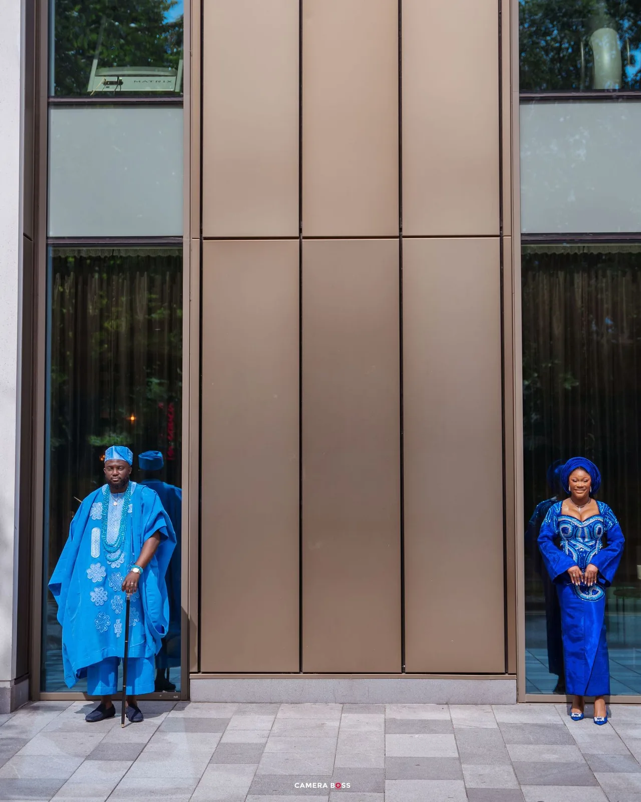 Two people in coordinating blue traditional African outfits stand in reflective doorways of a modern building.