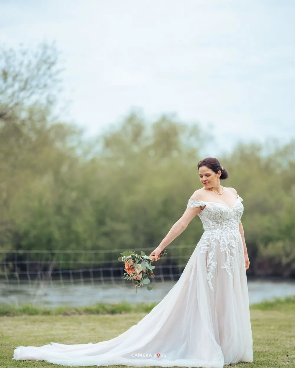 Bride in elegant off-shoulder lace wedding gown with long train poses in outdoor setting by waterfront.