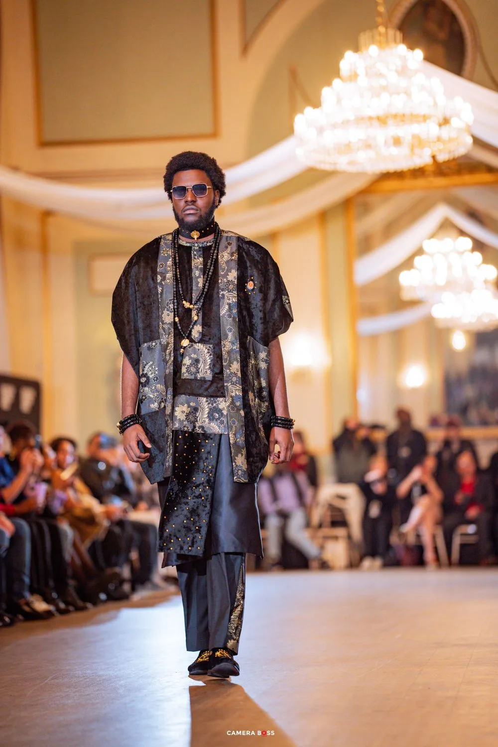 Model walks runway in luxurious black traditional African-inspired outfit with embroidery at elegant fashion show.