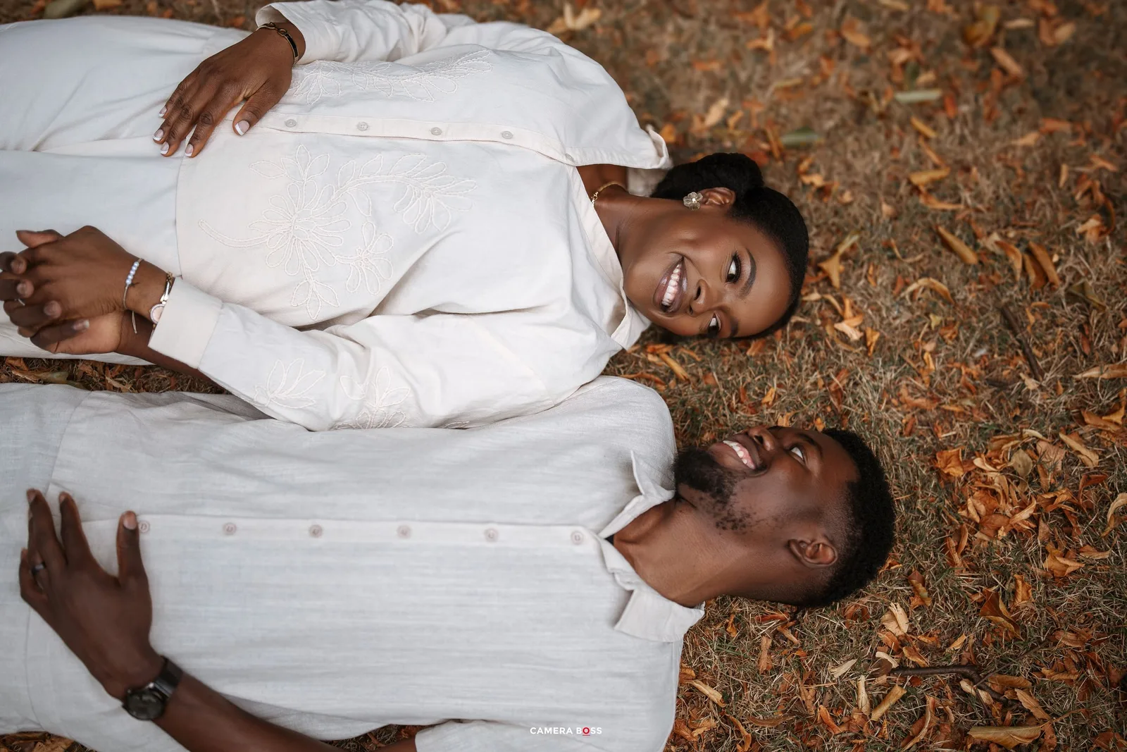 Two people in white clothing lying on autumn leaves while looking up and smiling.