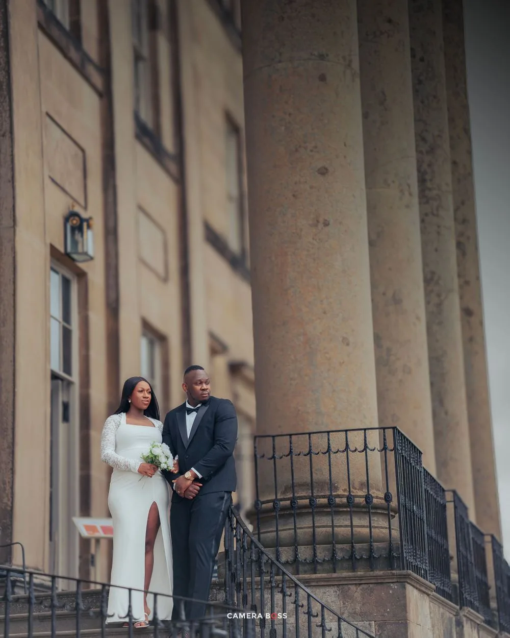 A couple in wedding attire poses elegantly on stone steps between classical columns at a historic building.