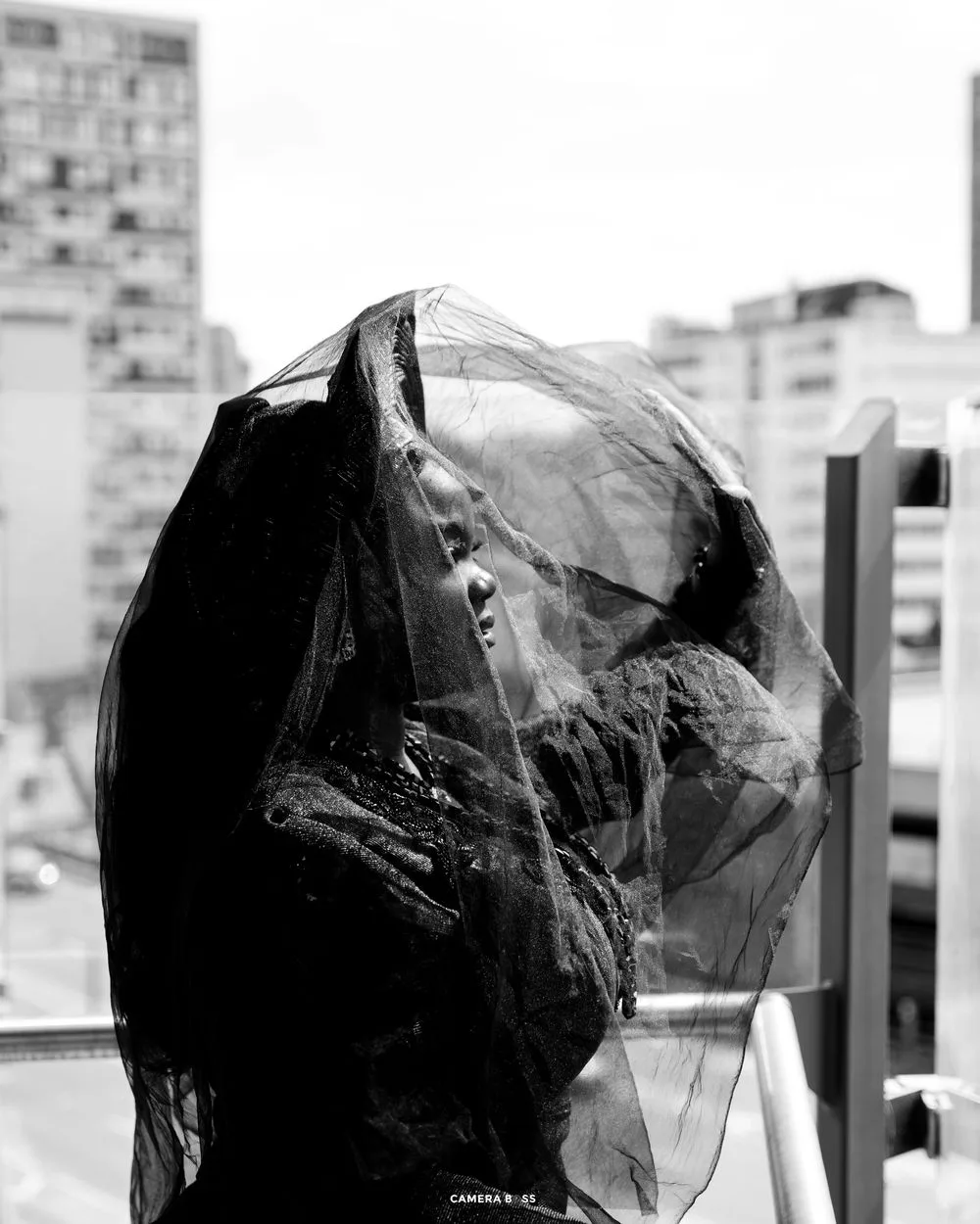 A figure covered in sheer fabric stands against an urban backdrop in dramatic black and white photography.