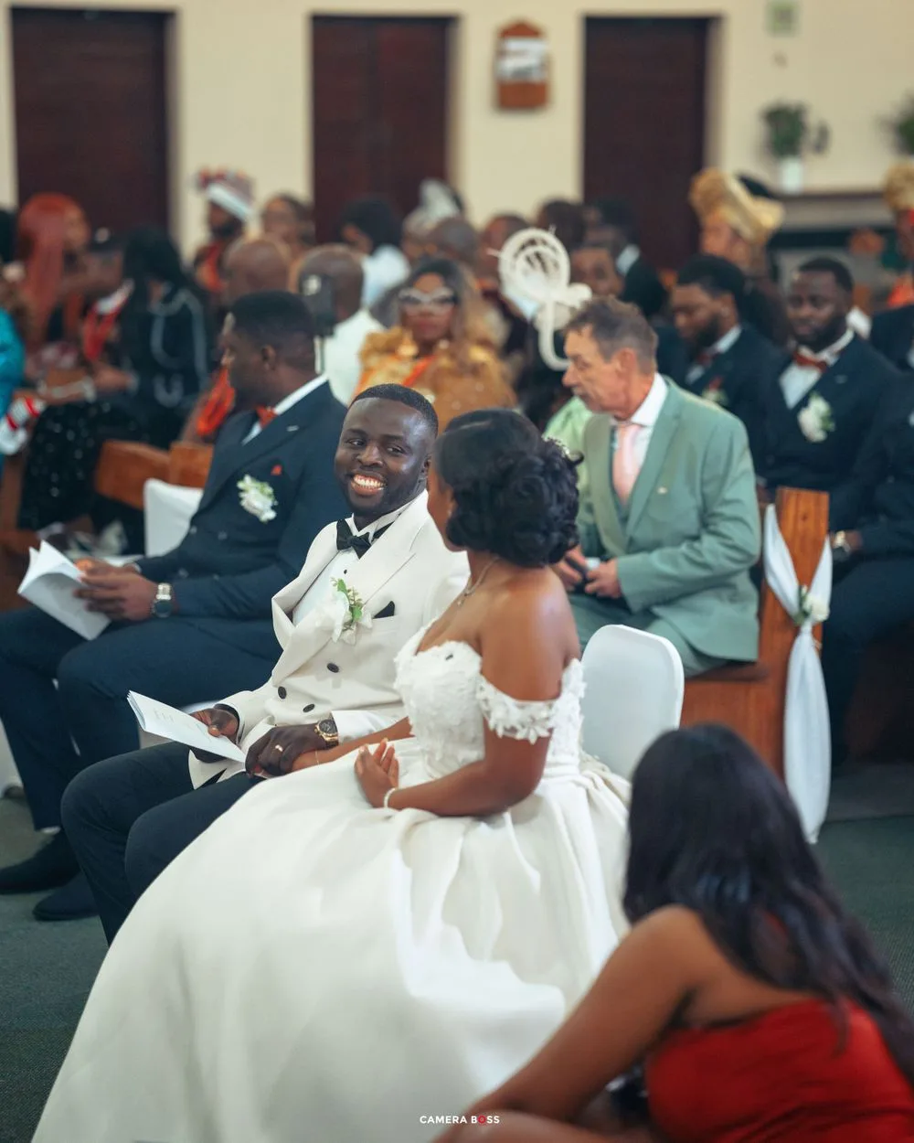 A newlywed couple shares a joyful moment seated among guests at their wedding ceremony in an indoor venue.