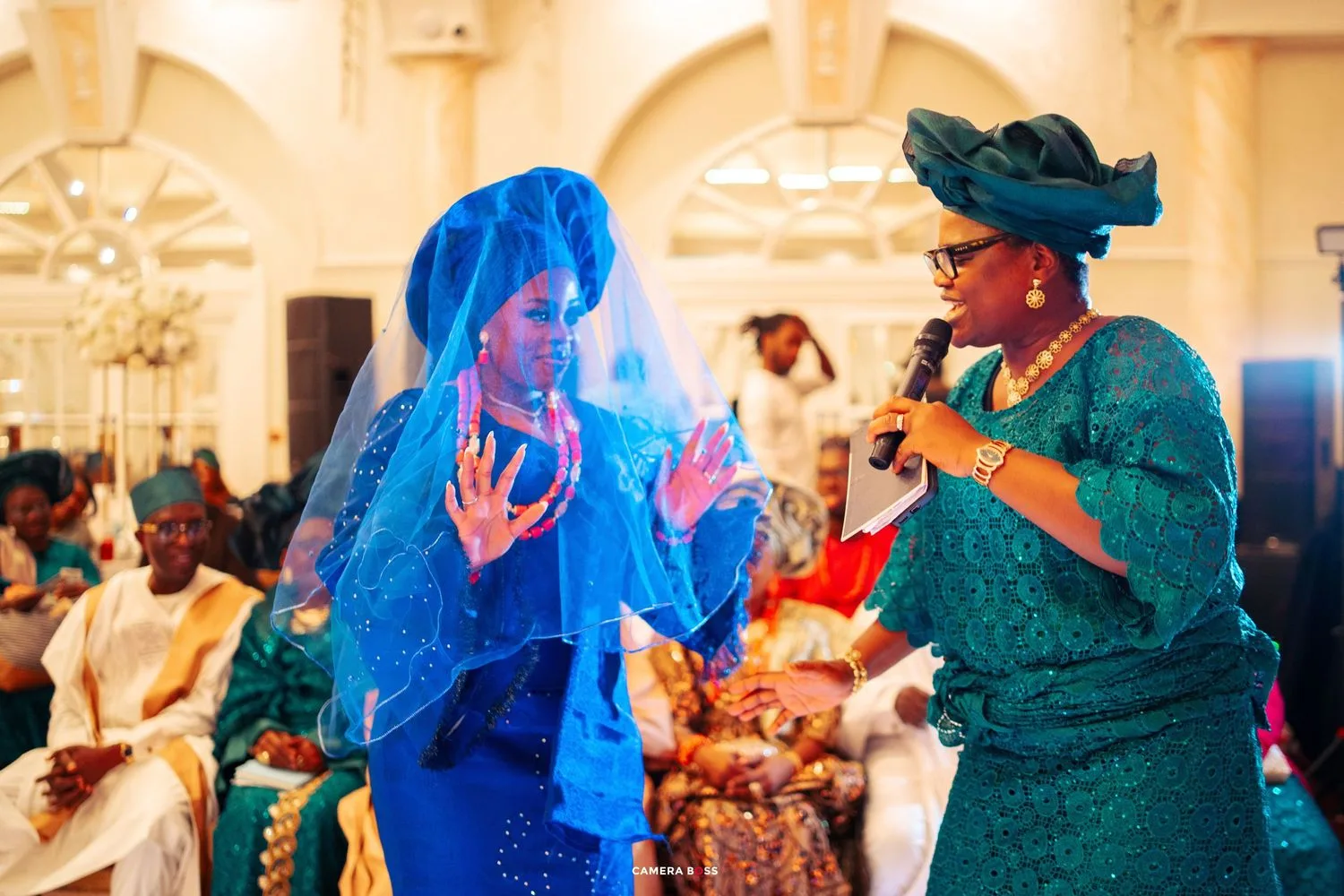 Two wedding guests in vibrant blue and green traditional attire dancing and celebrating at an elegant reception venue.